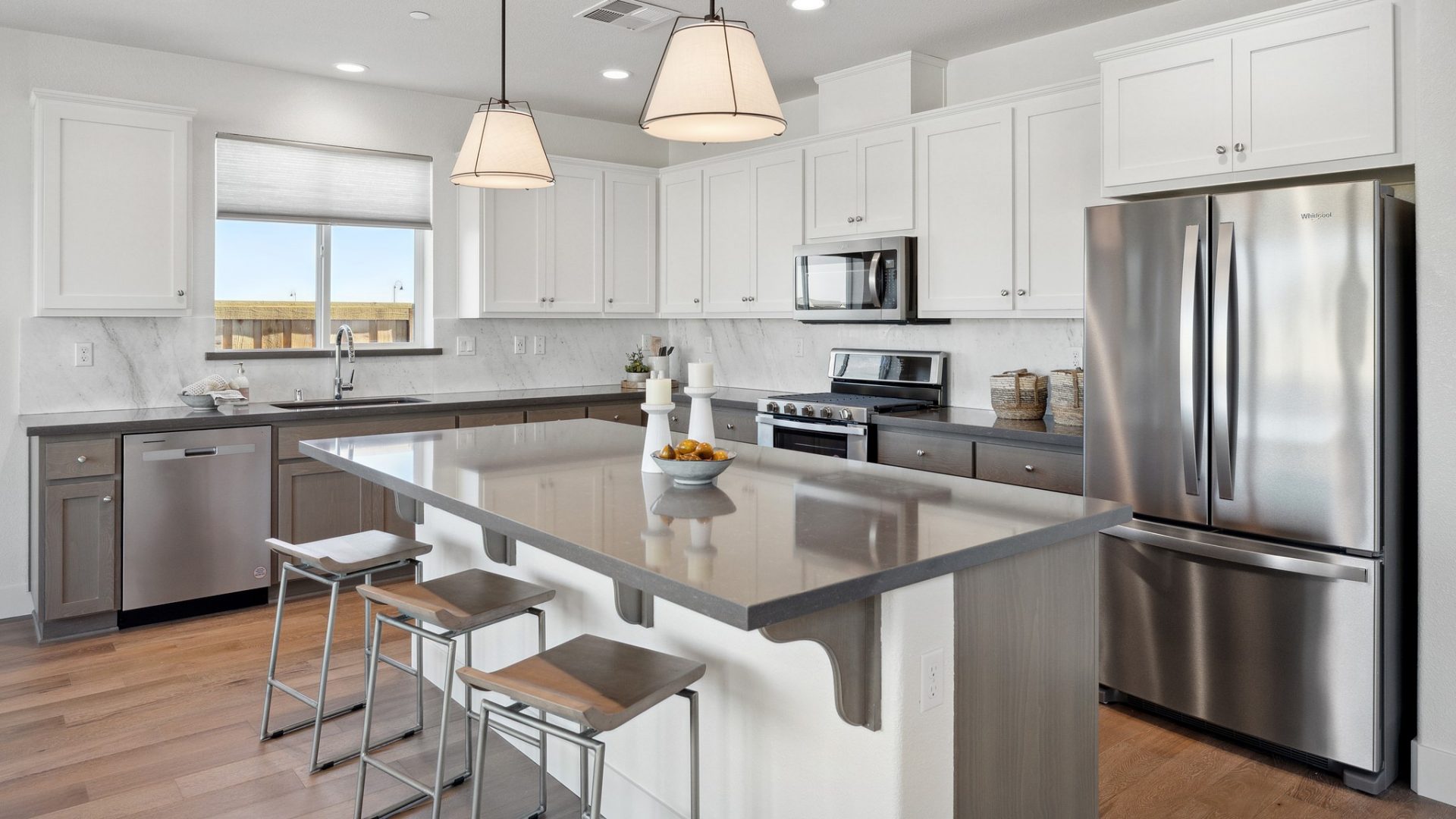 Open kitchen with hardwood floors, white beech wood cabinets, dark quartz countertops, stainless steel appliances, kitchen island with 3 counter stools, 2 pendant lights over the island and a large window over the kitchen sink.