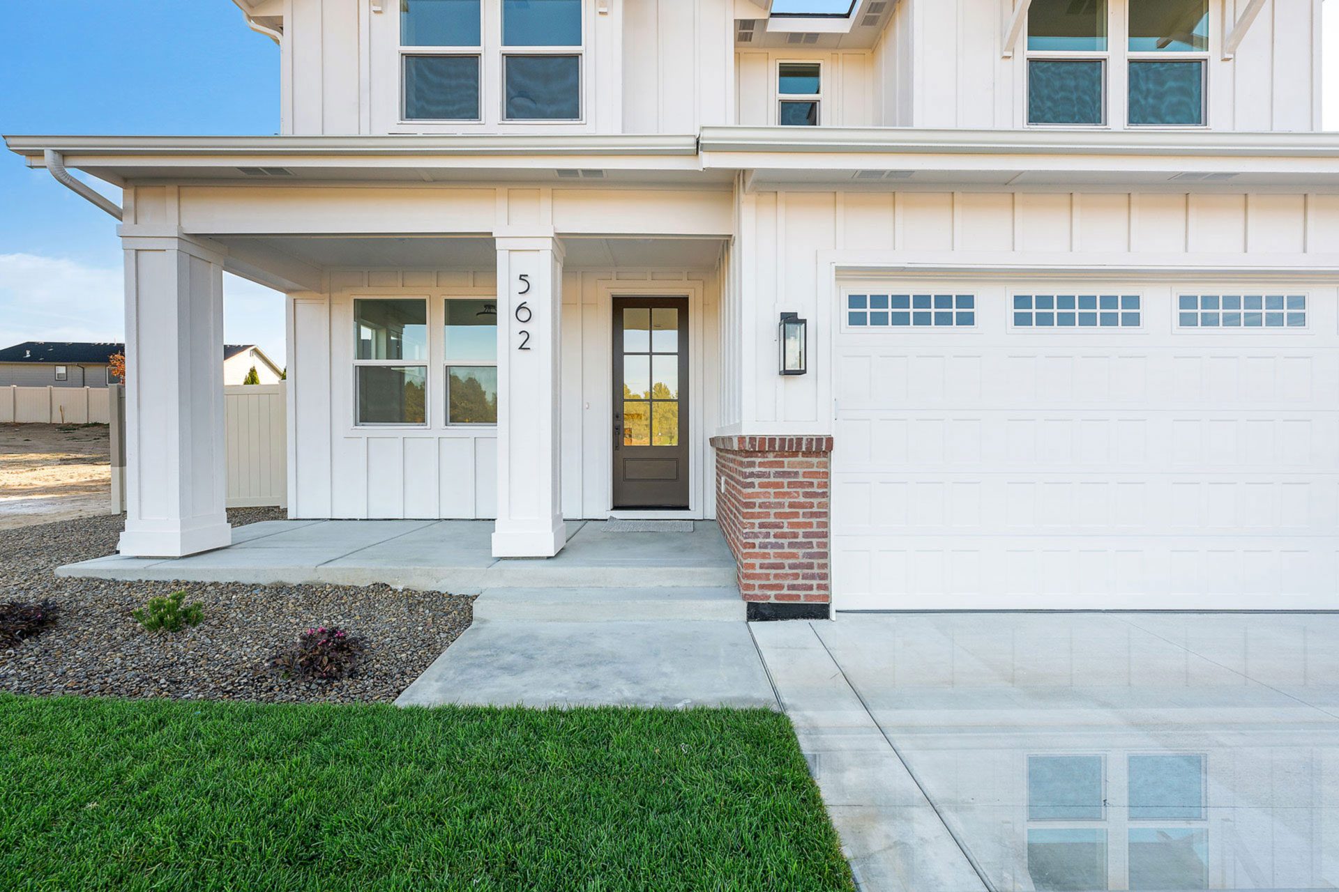 Lower half of front exterior elevation of farmhouse style 2-story home painted in white color tones, with covered porch, 2 car garage and grass landscaping.