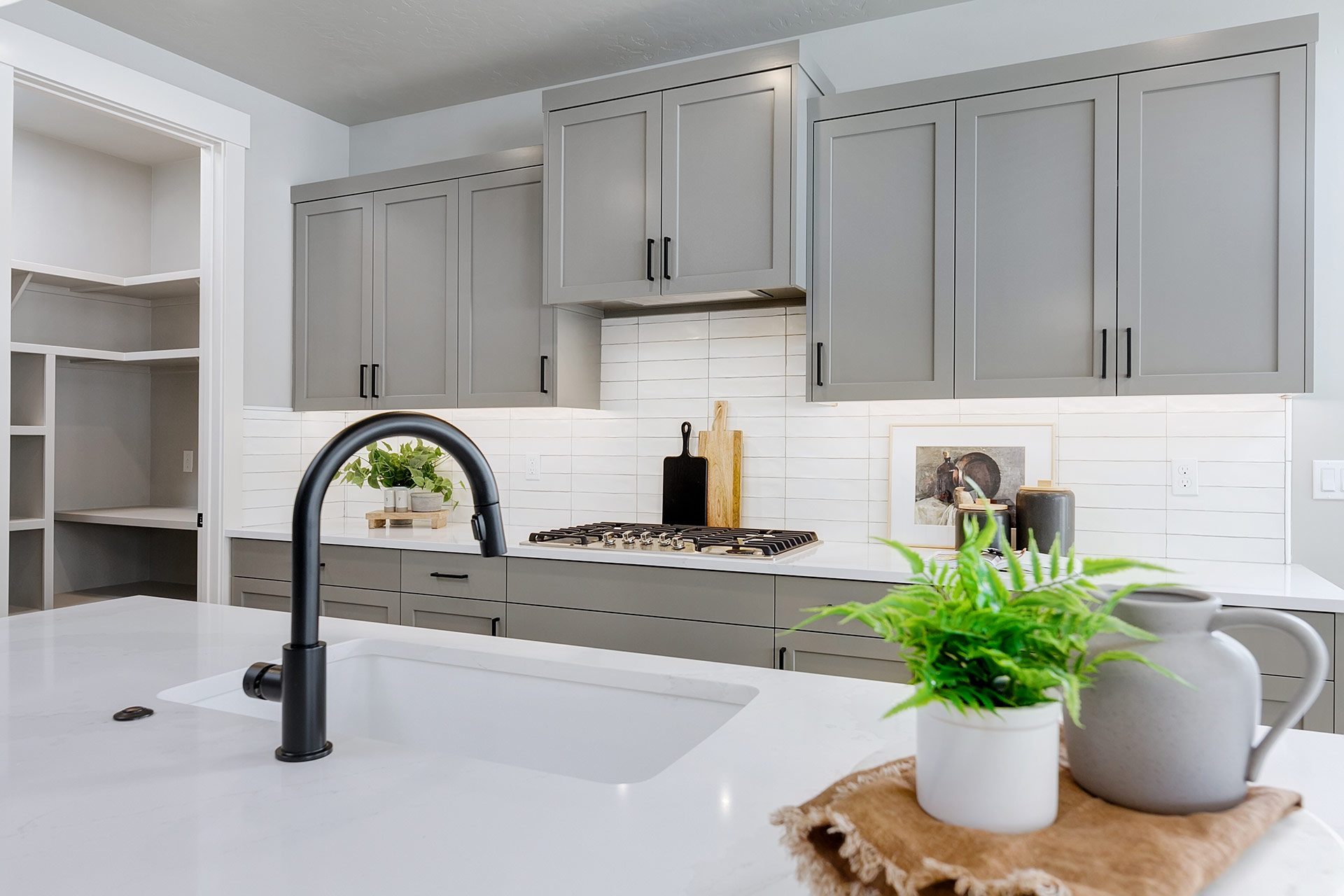 View of kitchen from the kitchen island with sink, black fixtures, white quartz countertops, grey beech wood cabinets and tiled backsplash.