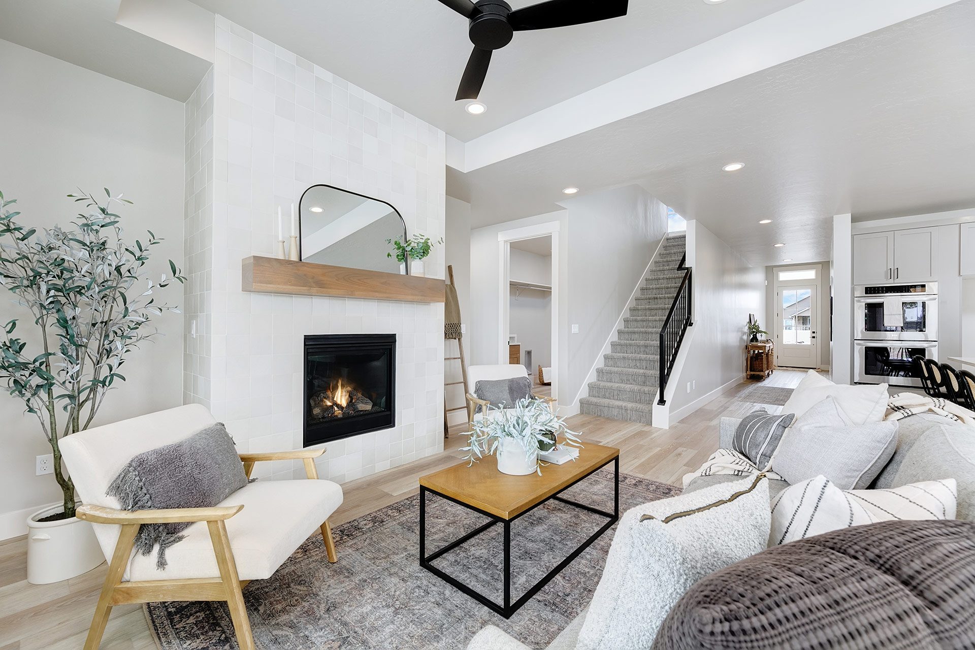 Open living room hardwood floors, 9 foot recessed ceiling and canned lights with a designer ceiling fan, tall mantel with electric fireplace, staged living room set and staircase to upstairs in the background.