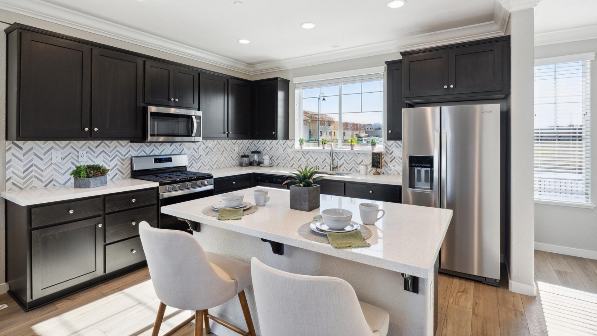 Open kitchen layout with hardwood floors, stainless steel appliances, dark beech wood cabinets, white quartz countertops, tiled backsplash, kitchen island with counter bar chairs, large kitchen window over the sink and 9 foot ceiling with canned lighting.