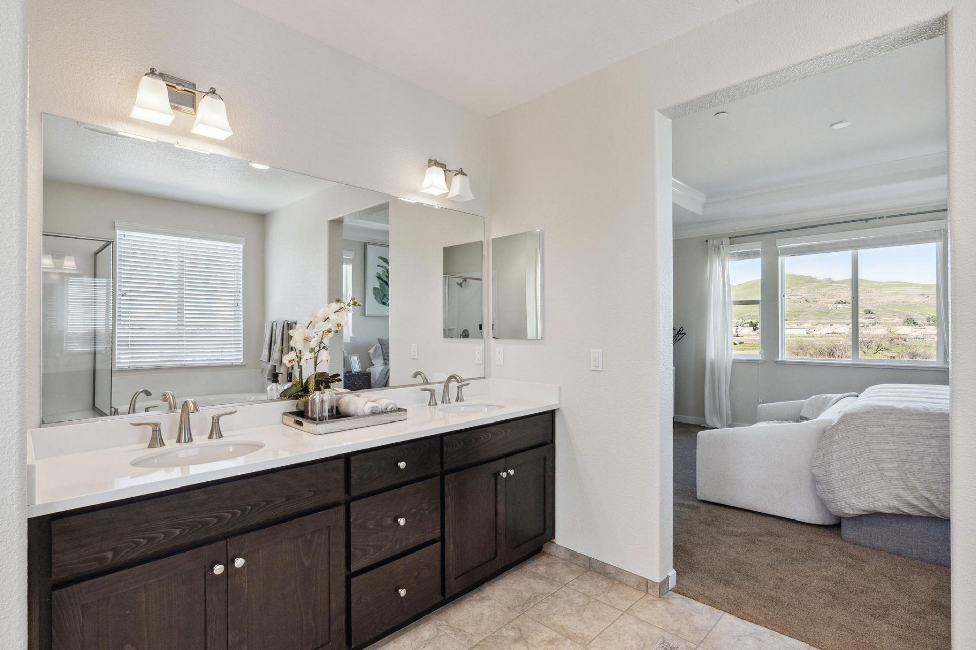 Primary bathroom with tiled flooring, dark beech wood dual sink vanity with white quartz countertop, sconce lighting above the vanity mirror, 9 foot ceiling with canned lights, and an 8 foot door entrance from the primary bedroom.