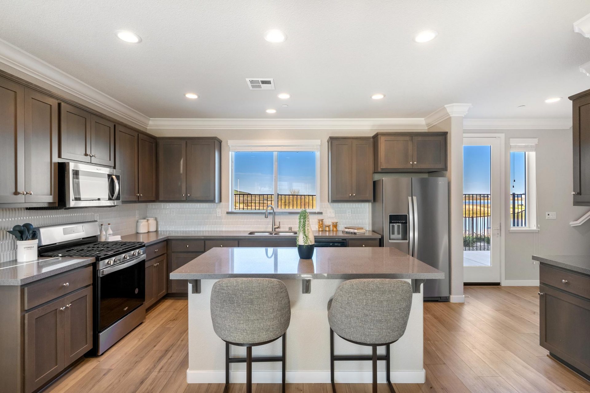Open concept kitchen with dark beech wood cabinets, grey quartz countertops, stainless steel appliances, hardwood floors, kitchen island with 2 counter bar chairs, and 9 foot ceilings with canned lighting.