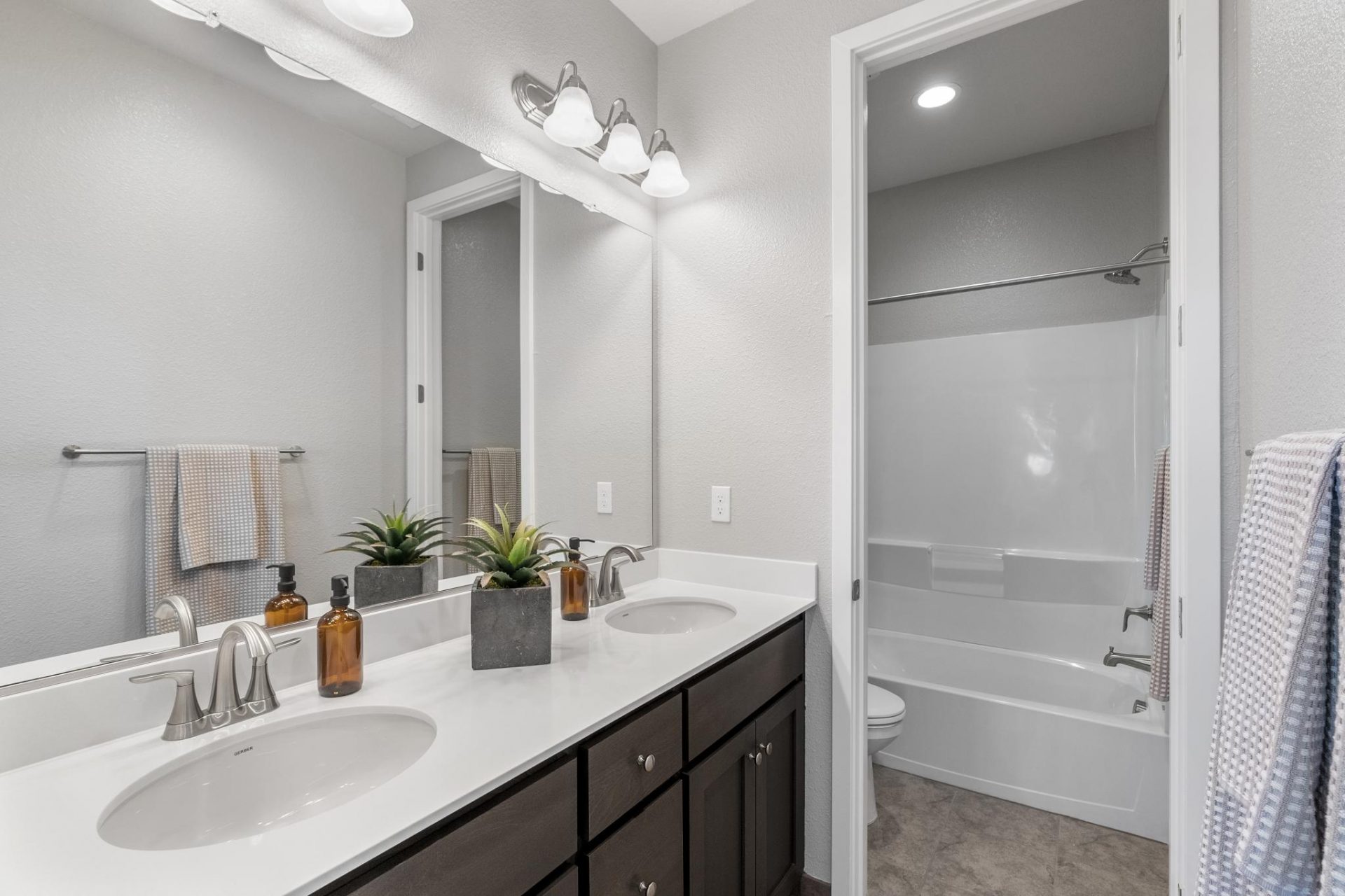 Dual sink vanity with dark beech wood cabinets, white quartz countertop, sconce lighting above the vanity mirror, 9 foot ceilings with canned lights, and an 8 foot door entrance to the shower with tub.