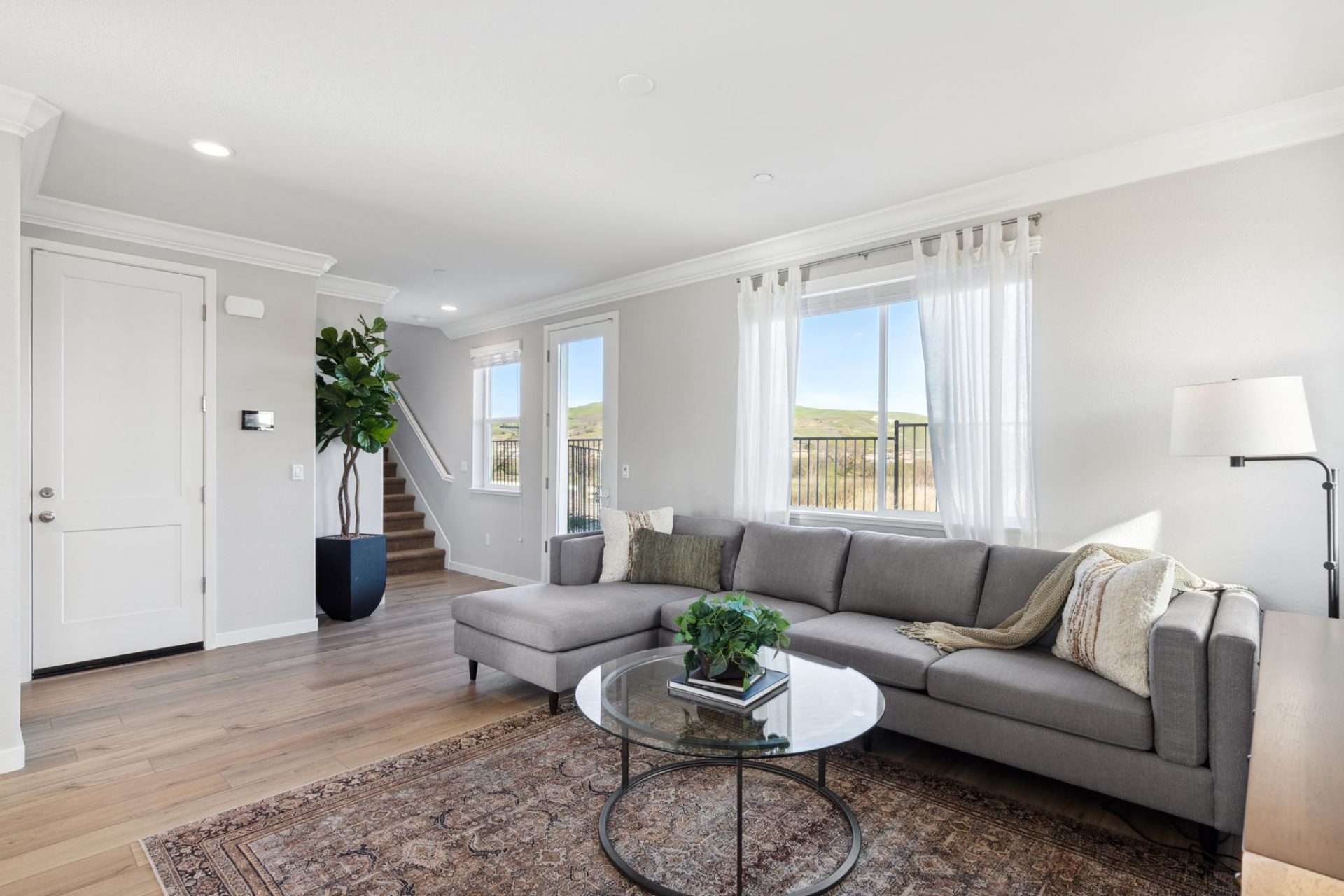 Open concept living room with hardwood floors, accent rug, glass coffee table, grey chaise couch, 3 large windows, 9 foot ceilings with canned lights and crown molding with 8 foot doors to the front entranceway.