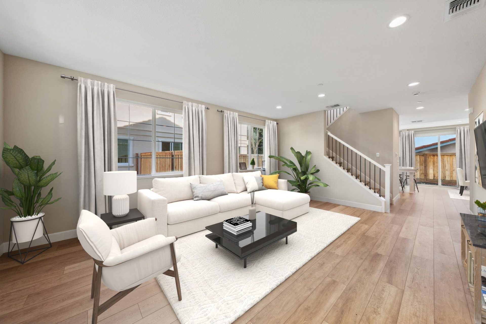 View of a virtually furnished room with brown wood texture flooring, a light colored sectional couch and chair, a dark coffee table and light colored area rug. Behind the couch are two windows with drapes, there are potted plants in two corners of the roo