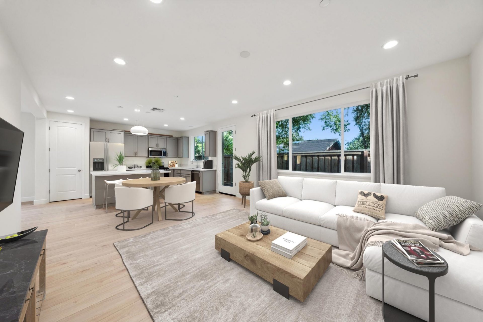 A living, dining and kitchen area open space virtually furnished with a white sectional couch and a square wooden coffee table,  in the dining area a light wooden round table with white chairs, kitchen in the background with white countertops and gray cab