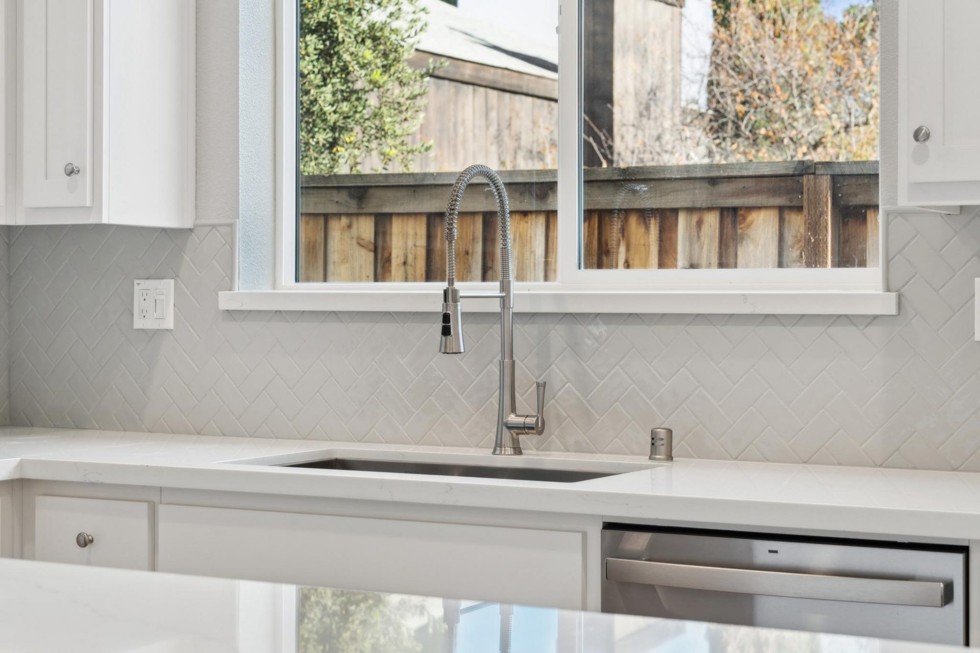 Photo of a kitchen sink with a brushed nickel faucet, tile backsplash, white countertops and a window over the sink.