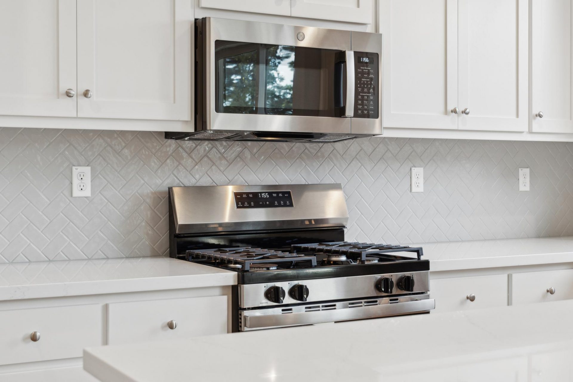 Photo of a kitchen with a stainless steel gas burner oven and stainless steel microwave above, countertop to cabinet tile backsplash in a herringbone pattern  and white cabinets.