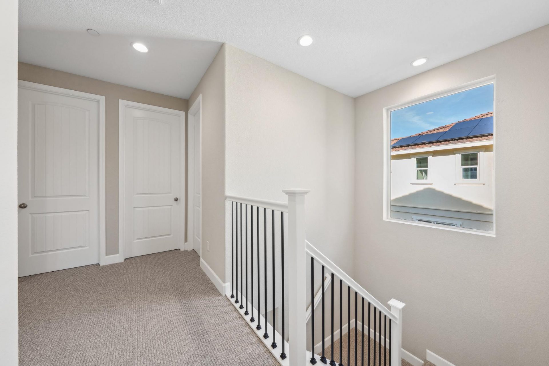 Photo of the top landing of a staircase with gray carpet, white doors to rooms on a far wall and to the left the staircase with white handrails and dark colored balusters and a window.