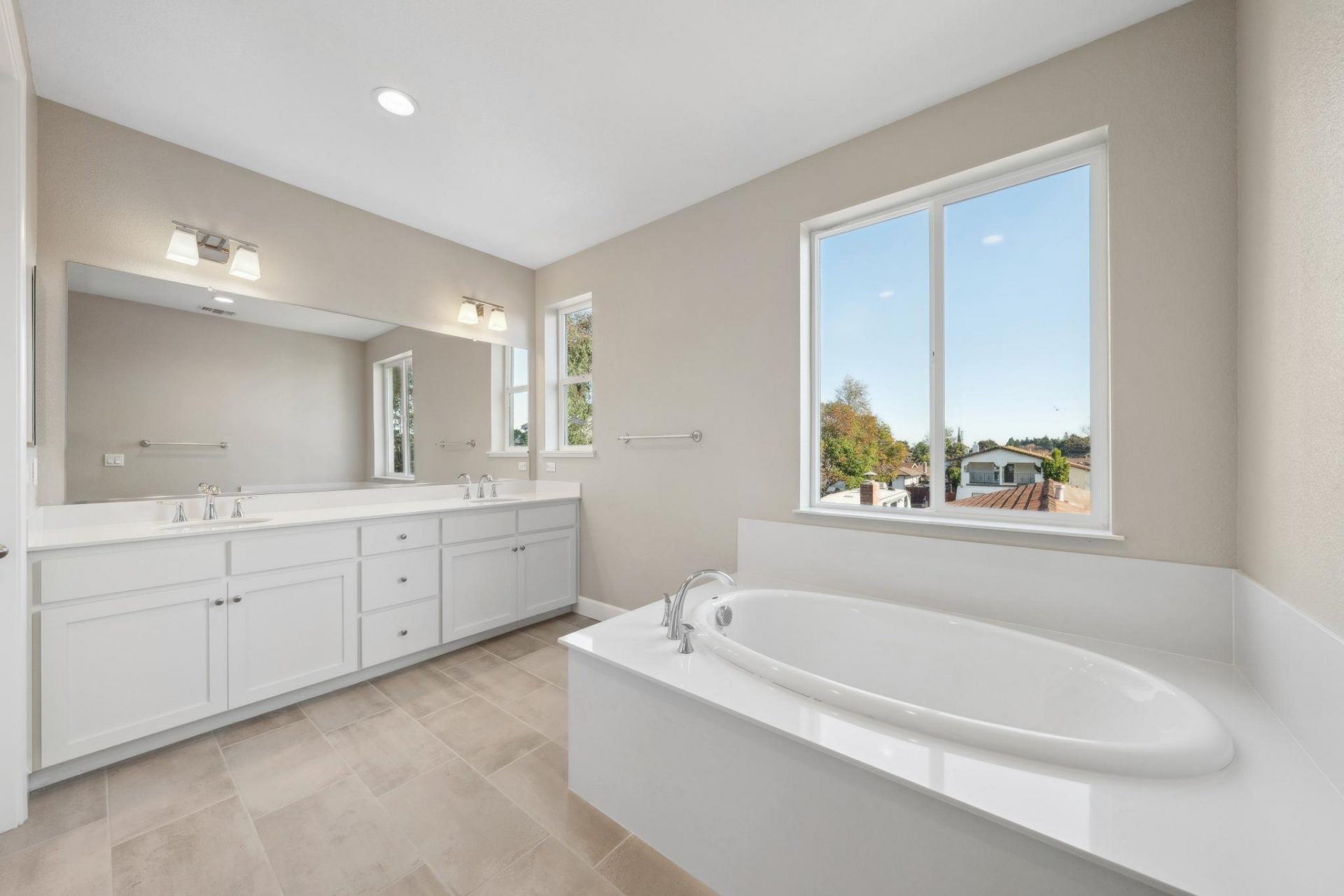 Photo of a bathroom with neutral toned floor tile, a white porcelain inlay tub with a window above it, a dual sink vanity with chrome fixtures, a mirror spanning the length of the vanity and two light fixtures above.