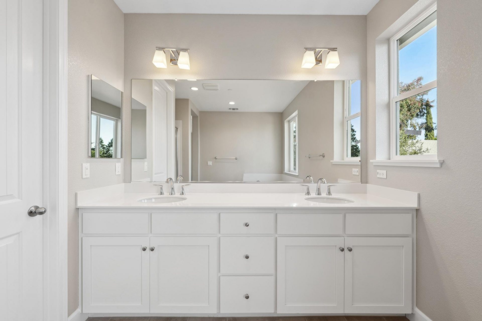 A double sink vanity with light color cabinets and countertop, chrome fixtures and a window that spans the length of the vanity, with two light fixtures above and a window to the left.