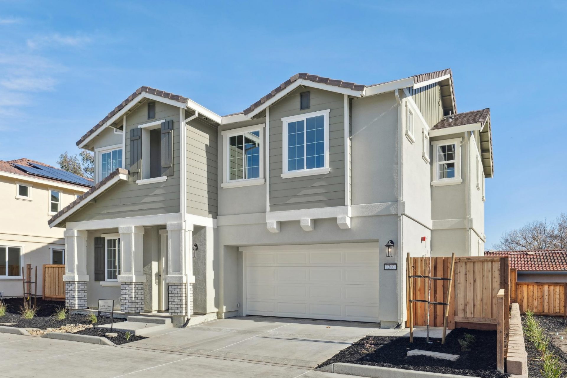 An angled view of the exterior of a modern two story home, with tile roofing, stucco and wood accents, with a modern color scheme, light colored garage door, three pillars at the entry with stone accents.