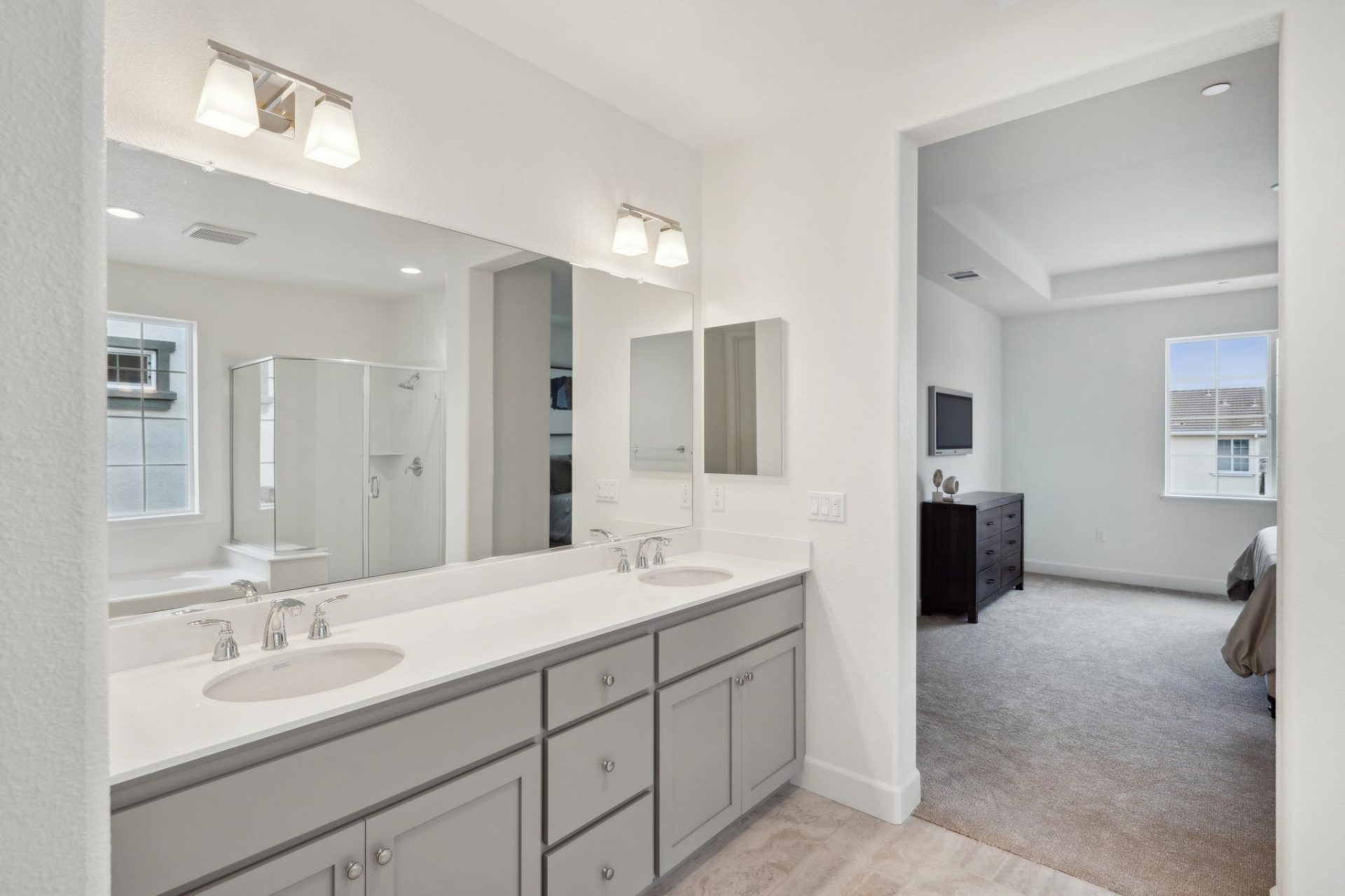 A bathroom with a dual sink vanity, with gray cabinets, white countertop, and chrome faucets. A long mirror and two light fixtures located above the vanity. To the right is a bedroom.