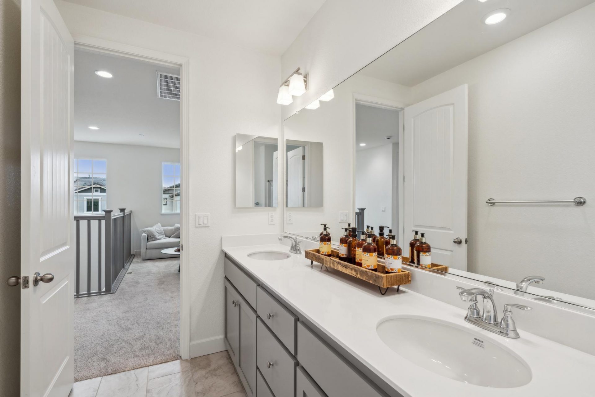 A bathroom with a dual sink vanity, with gray cabinets, white countertop, and chrome faucets. A long mirror and a light fixture located above the vanity. Straight ahead is a loft/seating area.