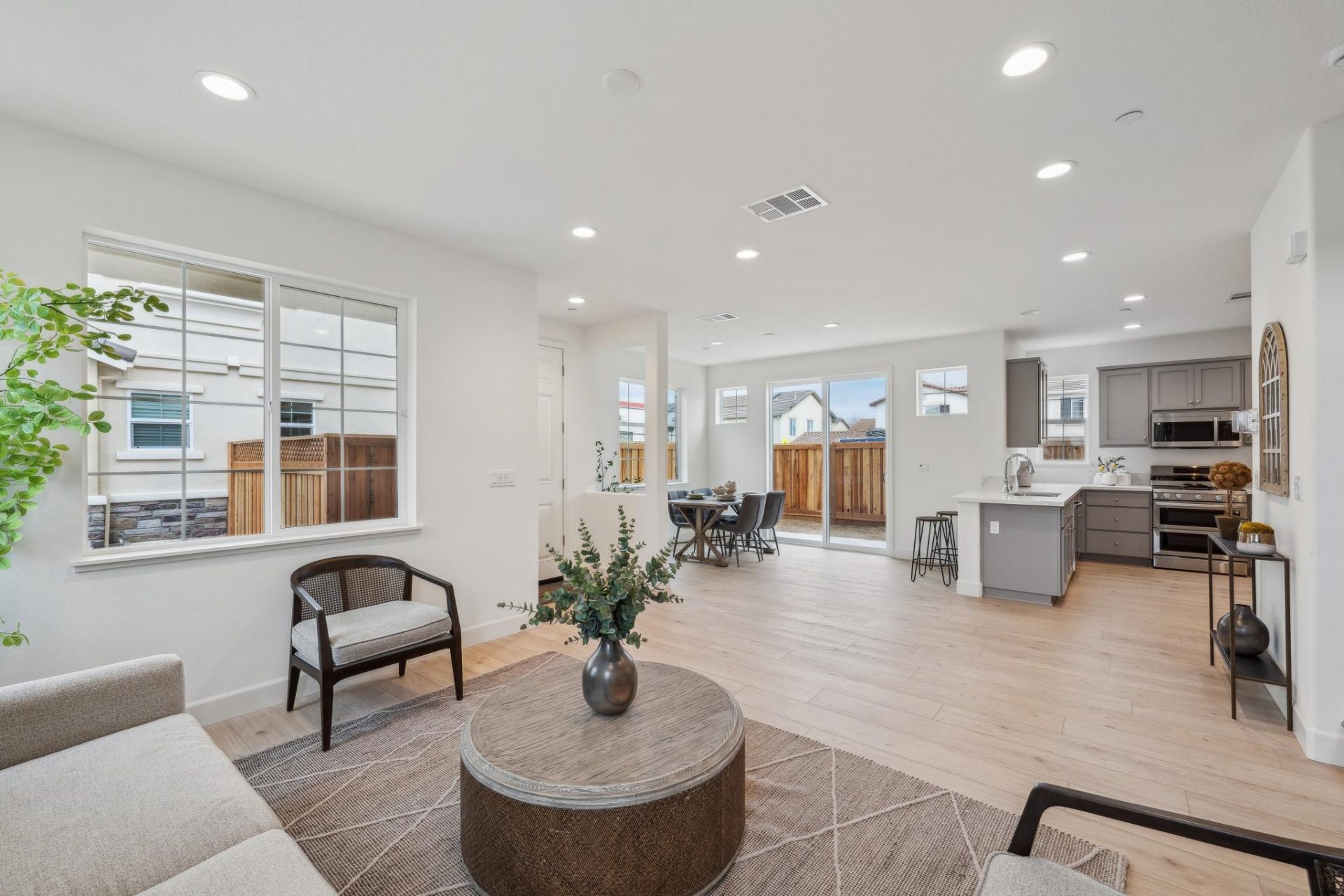 A partial view of a staged seating area with a dining area in the distance to the left and a kitchen to the distance on the right. The open space area has light colored wood flooring, can lights in the ceiling and lots of windows.