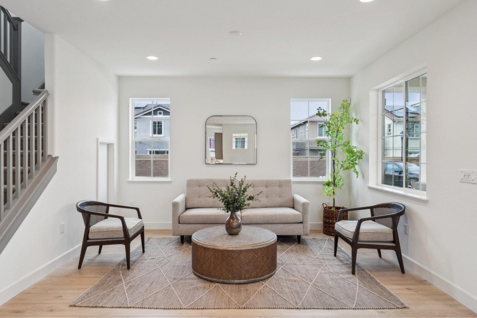 A living area furnished with a light color couch, two seating chairs, a round coffee table, area rug and a potted plant. Several windows in the space create a bright area.