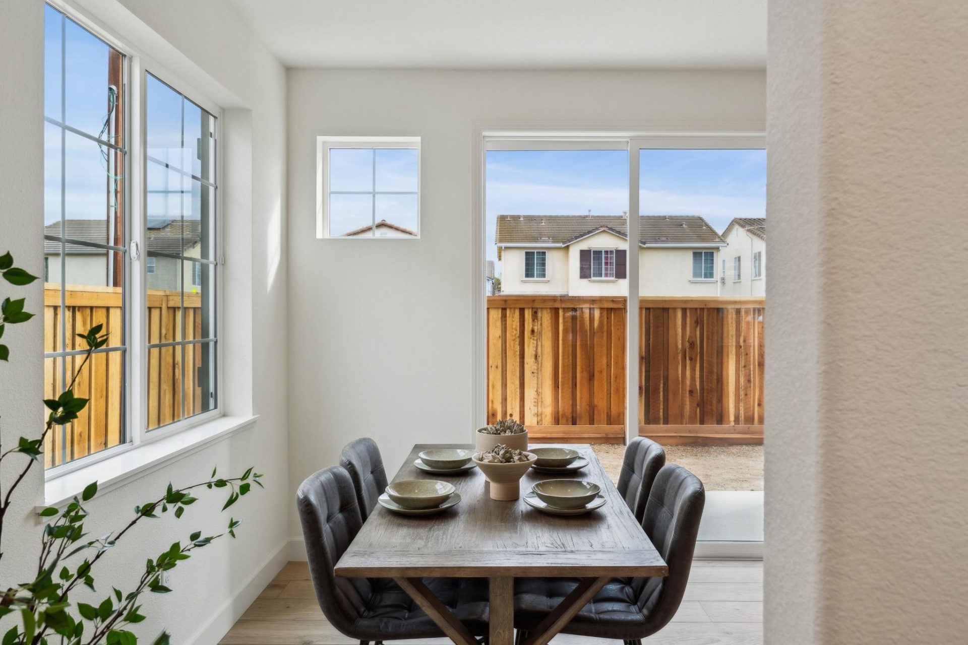 A furnished dining area with a rectangular table with four chairs and table setting. Windows to the left and a sliding glass door is seen slightly right behind the dining table.