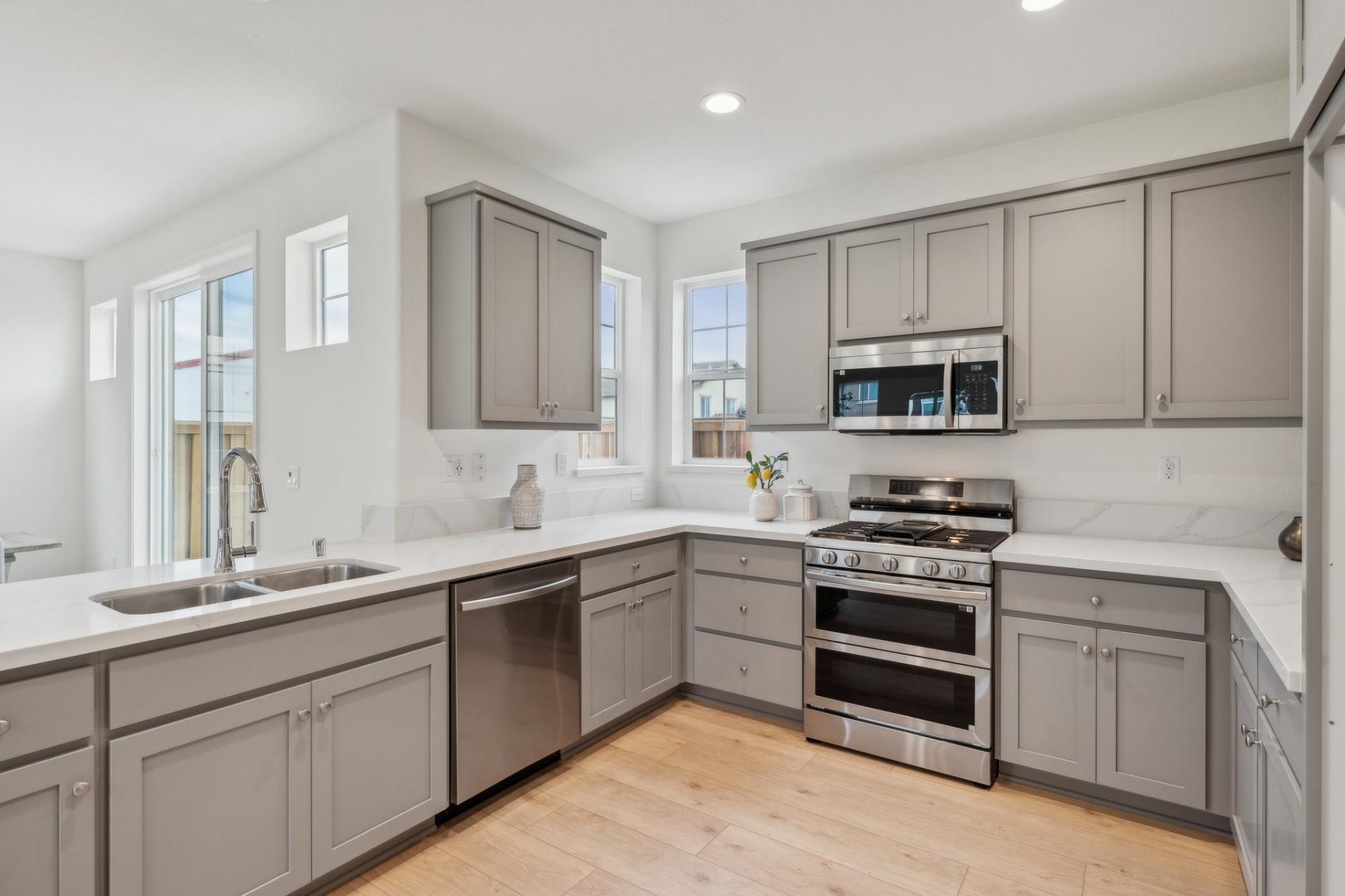 A modern kitchen with gray color upper and lower cabinets, a light colored countertop with stainless steel appliances, light brown wood flooring, light colored walls and two windows brightening the space.