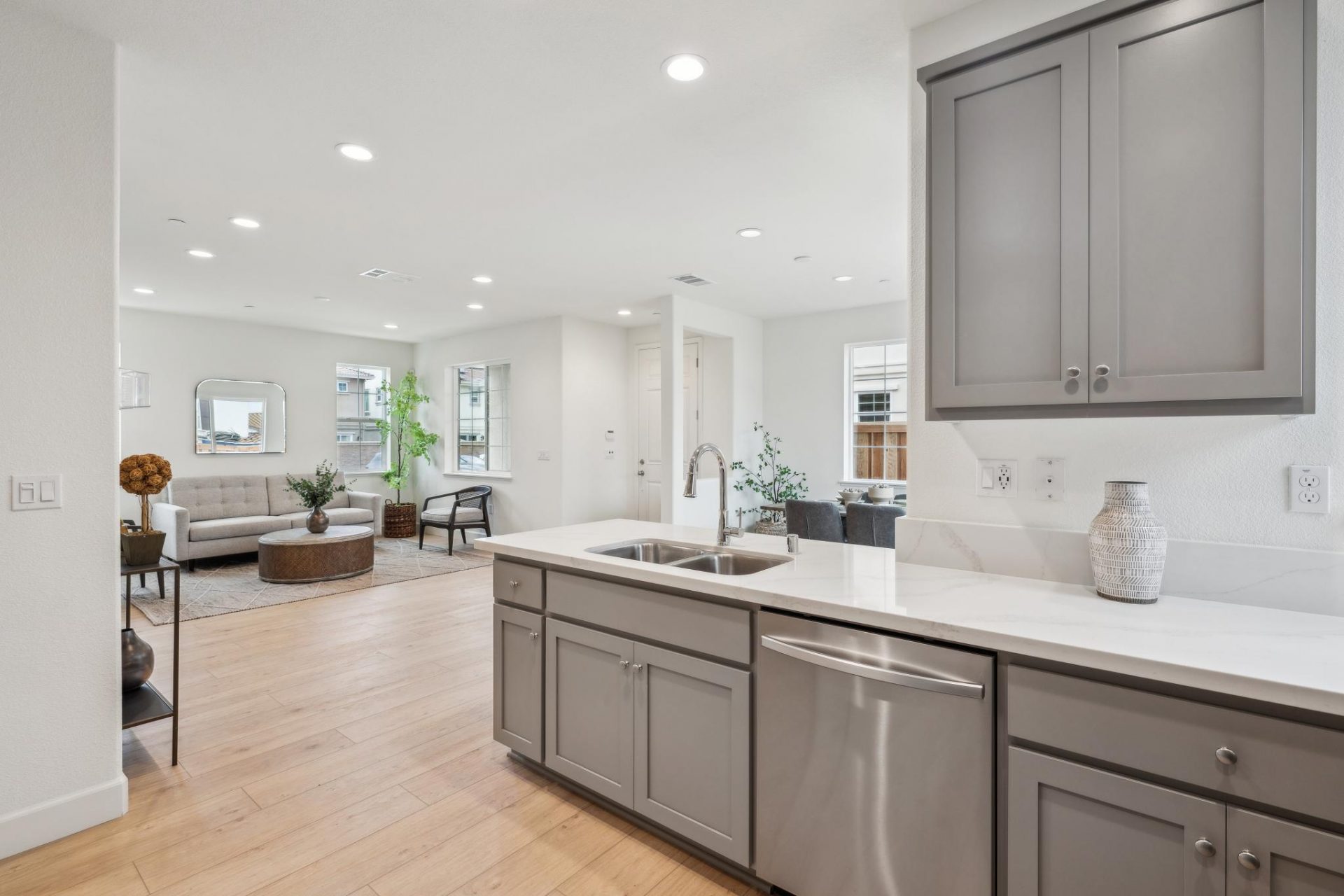 A peninsula kitchen countertop with cabinets and a kitchen sink with a dual sides and a chrome faucet. In the background a living/seating area to the left and a dining area to the right.