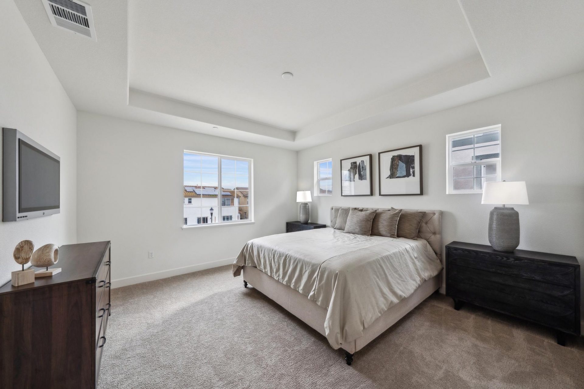 A bedroom with high ceilings, three windows, medium brown carpet, and furnishings including a bed, two nightstands with lamps, wall art, a dresser and a tv.