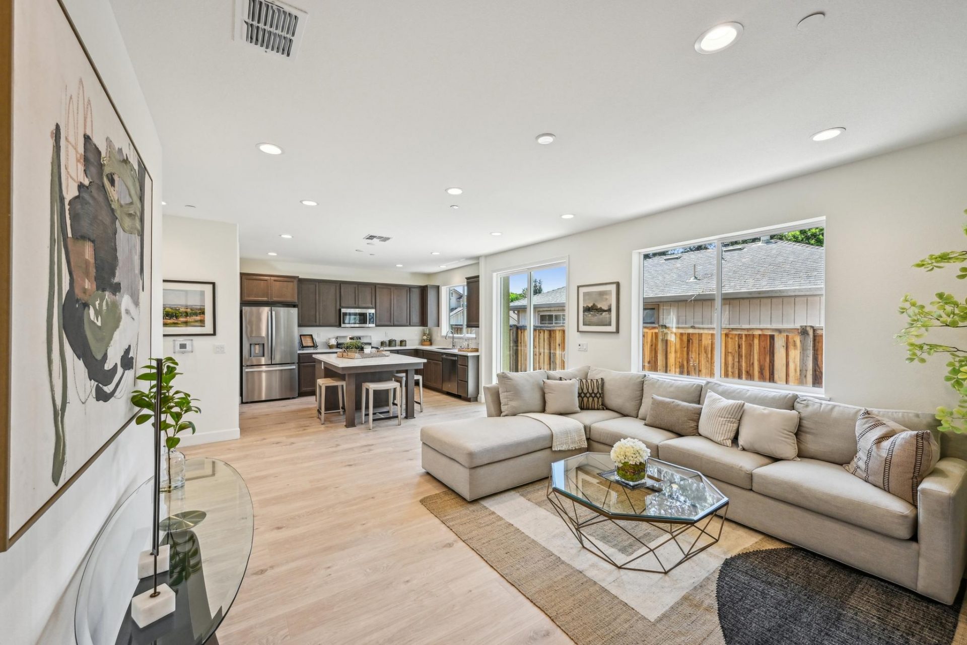 Open concept living room with hardwood floors, accent rug, glass coffee table, 9 foot ceilings with canned lighting, chaise couch and kitchen in the background.