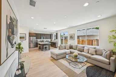 Open concept living room with hardwood floors, accent rug, glass coffee table, 9 foot ceilings with canned lighting, chaise couch and kitchen in the background.