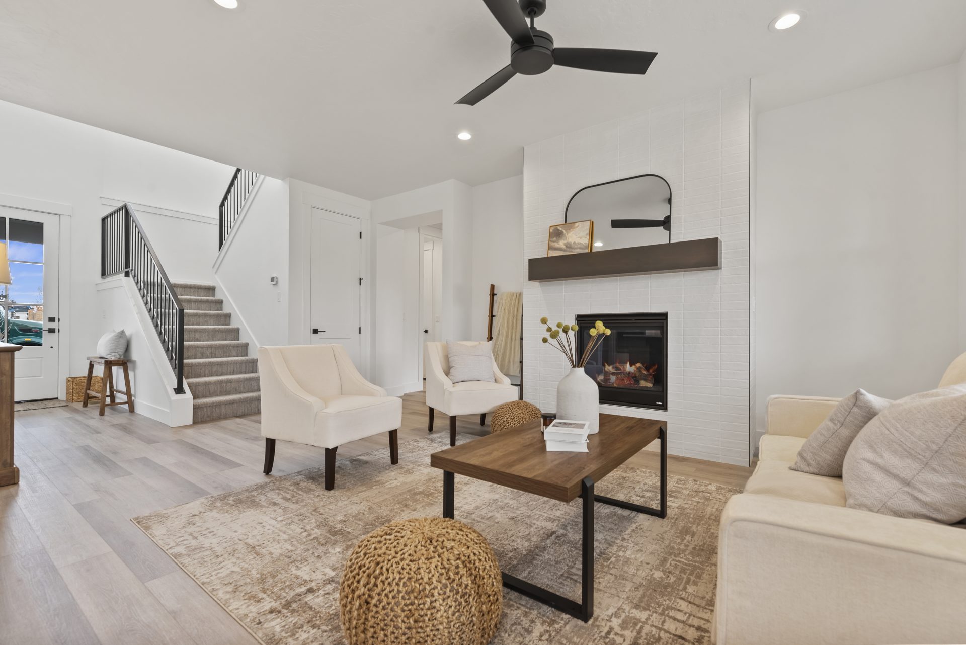 Living room with hardwood floors, 9 foot ceilings with canned alights and ceiling fan, tall mantel with electric fireplace, staircase to upstairs in the background.