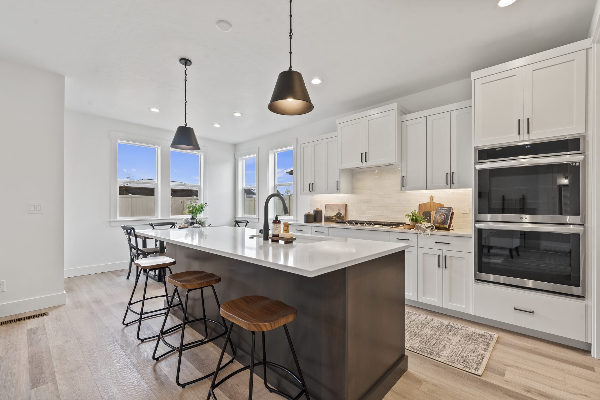 Open kitchen with hardwood floors, white beech wood cabinets, white quartz countertops, kitchen island with 3 barstools, stainless steel appliances, 4 windows, and 9 foot ceilings with canned lights and 2 pendants over the island.