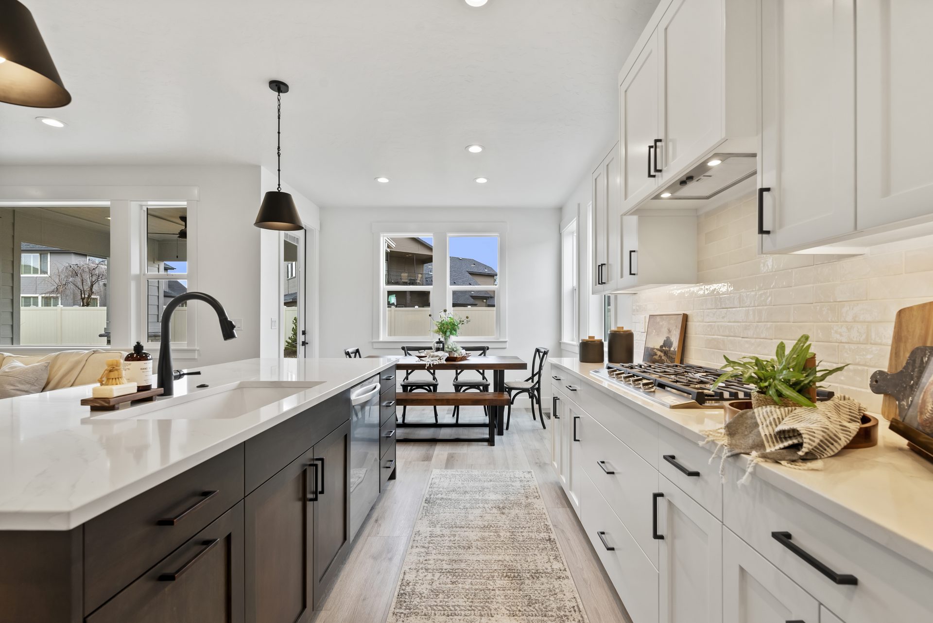 Open kitchen with hardwood floors, white beechwood cabinets, stainless steel stovetop, kitchen island with sink, white quartz countertops, 2 pendant lights over island, looking towards many open windows with natural light.