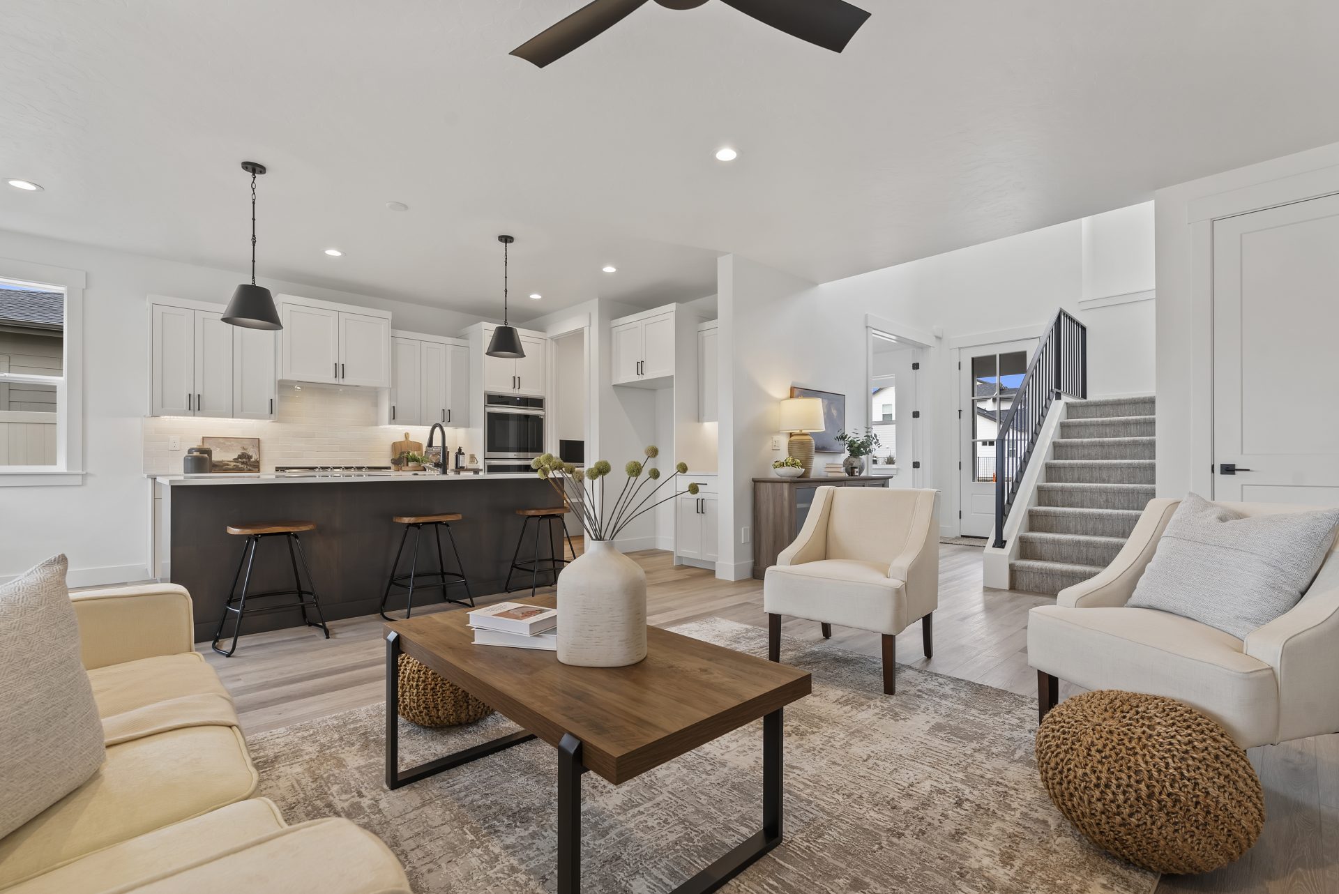 Open concept living room and kitchen with staircase to upstairs in the background, hardwood floors, 9 foot ceilings with canned lights, ceiling fan in living room, pendant lights over kitchen island.