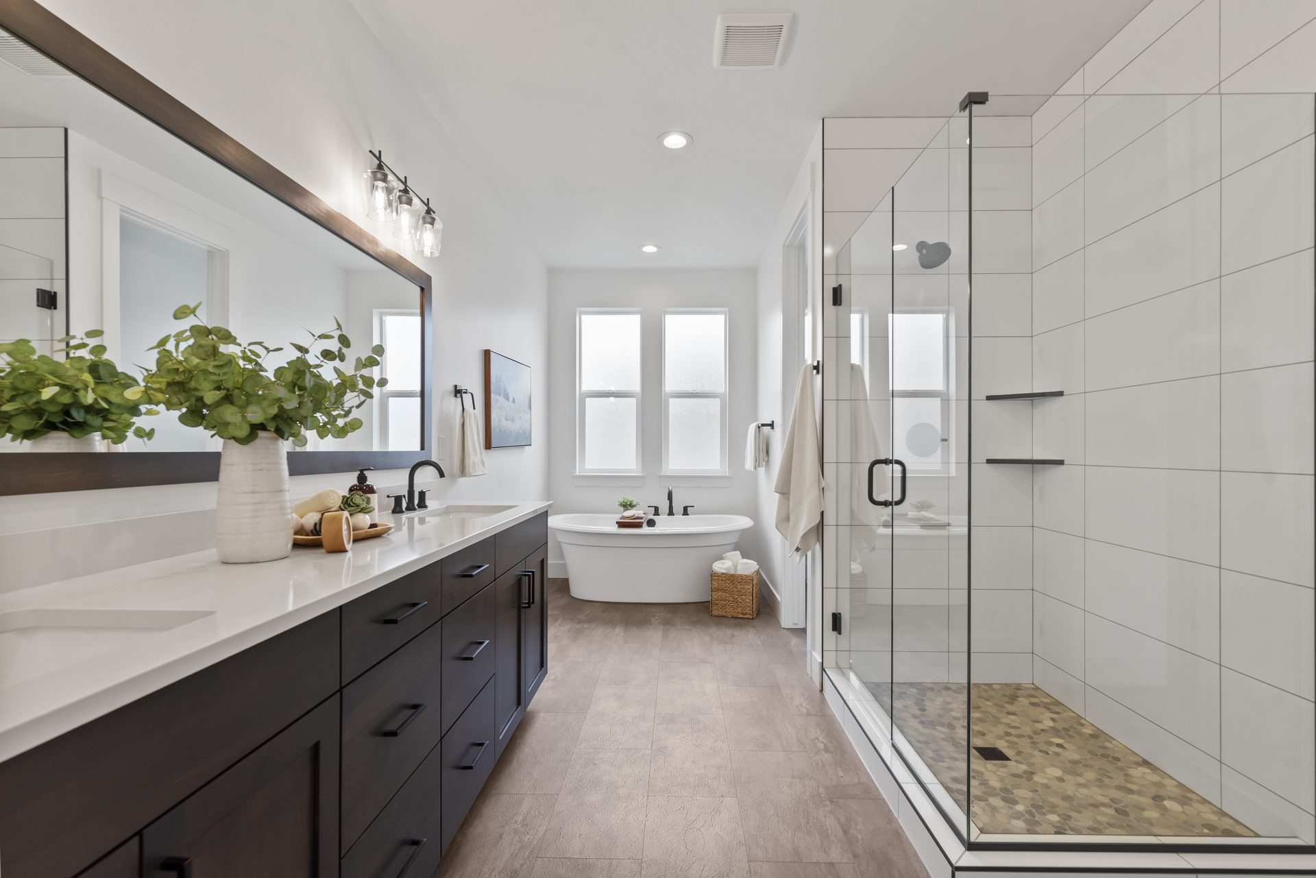 Tiled primary bathroom with dark beechwood vanity, dual sinks, designer sconces over mirror, tiled walk-in shower with glass doors, a soaking tub and 2 windows.