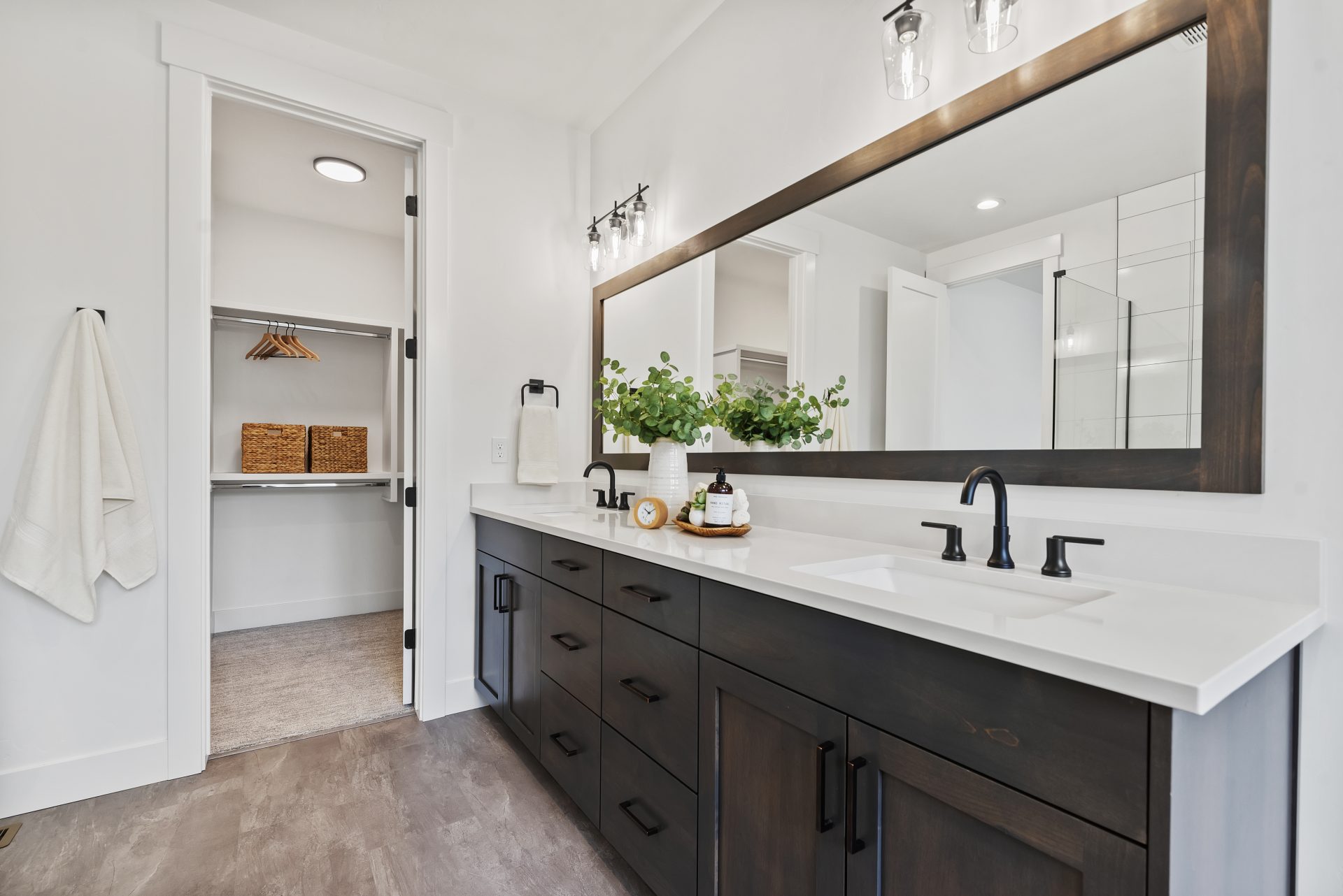 Tiled bathroom with dual sink vanity made of dark beechwood, white quartz countertops, designer sconces over the vanity mirror, black fixtures, 9 foot ceiling with canned lights and 8 foot doorway to the carpeted walk-in closet.