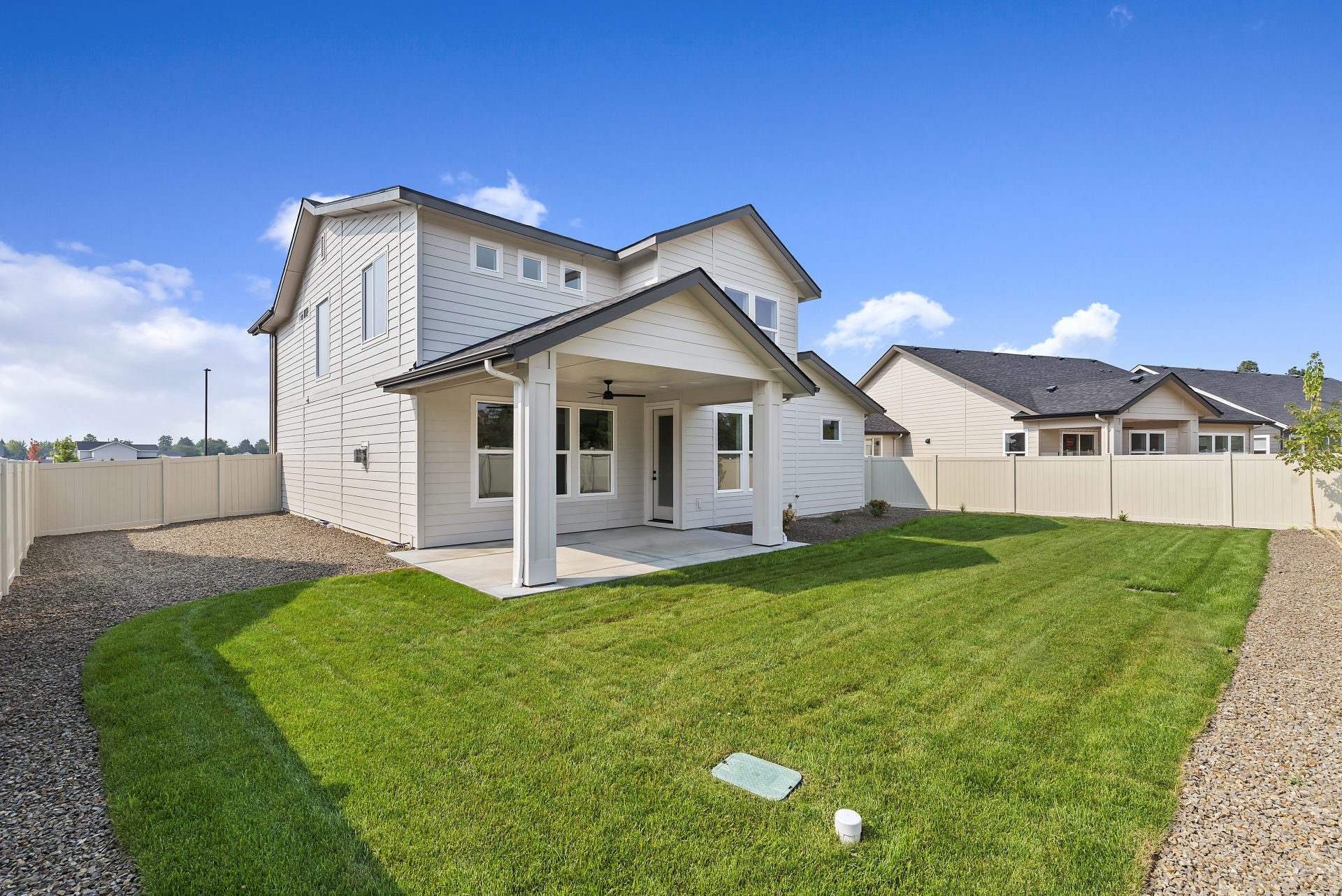 Backyard view of a modern two-story home with light siding, a covered patio, and a green lawn bordered by gravel and a light tan privacy fence, with the side of the house and multiple windows visible