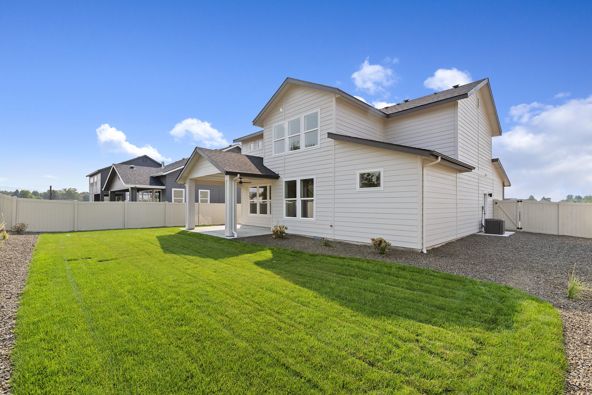 Backyard view of a modern two-story home with light siding, a covered patio, and a neatly landscaped lawn surrounded by a light tan privacy fence.