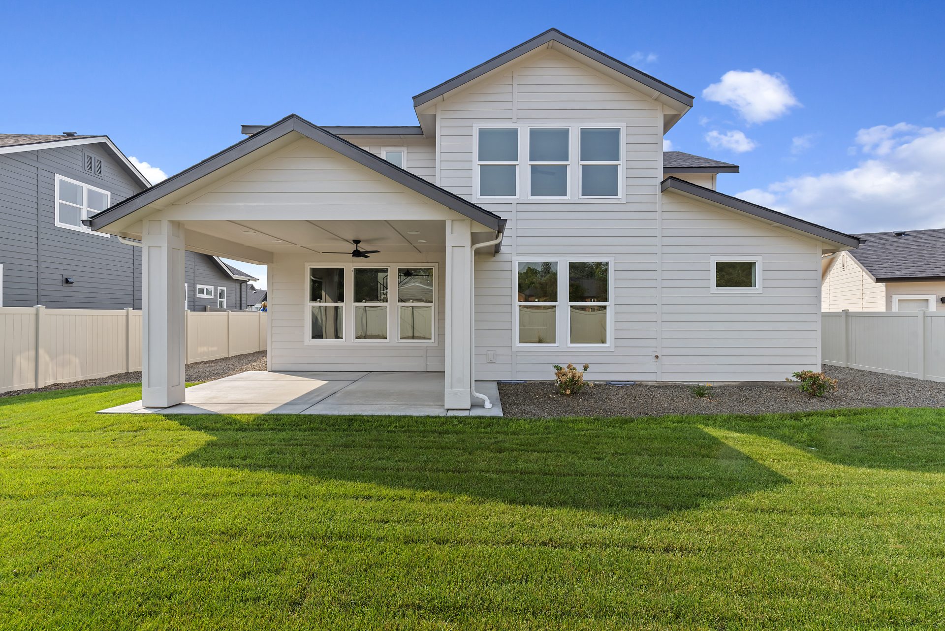 Backyard view of a modern two-story home with light siding, a covered patio, and a green lawn bordered by gravel and a light tan privacy fence, showing the patio ceiling fan and large rear windows.