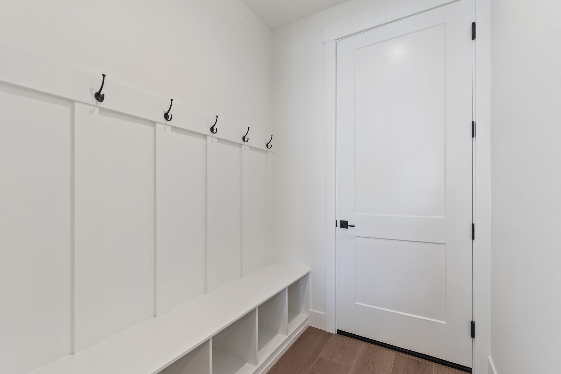 Bright mudroom featuring white paneled walls and a white door, with a built-in bench and cubby storage. The space has medium brown wood flooring, creating a clean and welcoming entryway.