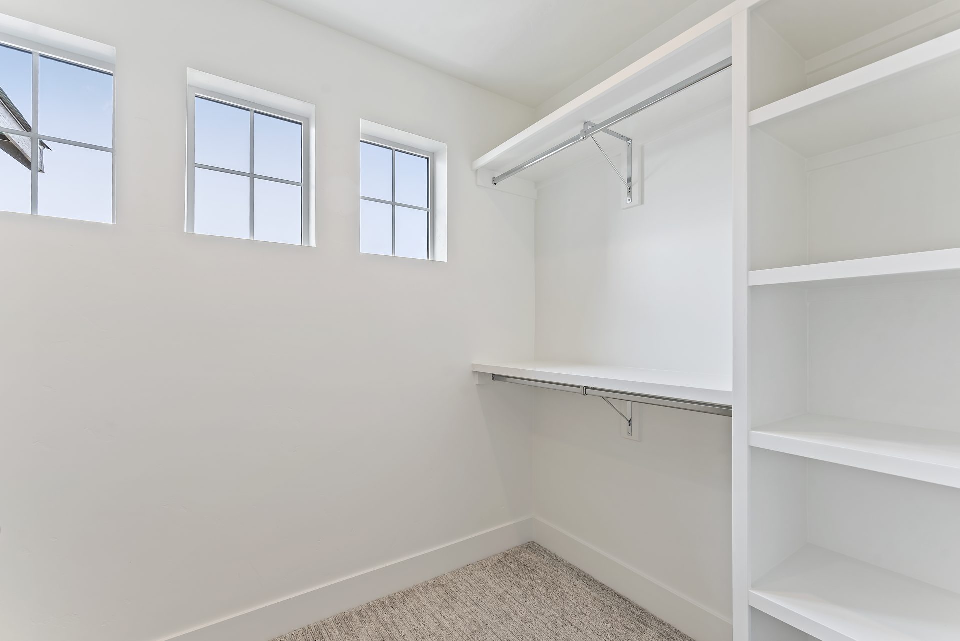 Walk-in closet featuring three small square windows, approximately 18 by 18 inches each, two-tiered hanging bars, four shelves for storage, and carpeted flooring