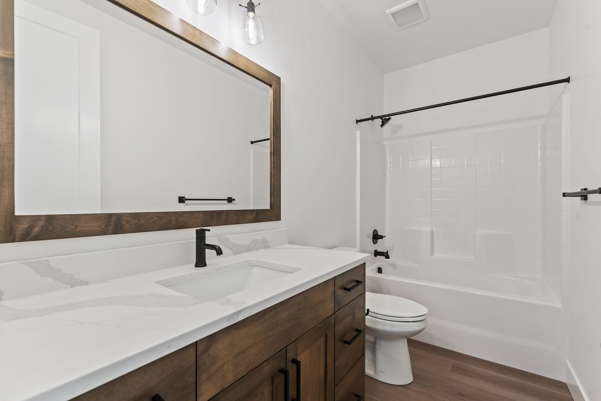 Bathroom featuring a wide stained brown vanity with white countertop, black faucet, and an equally wide mirror with matching brown frame. Includes a tub/shower unit with black finishes and a toilet, with medium brown wood flooring