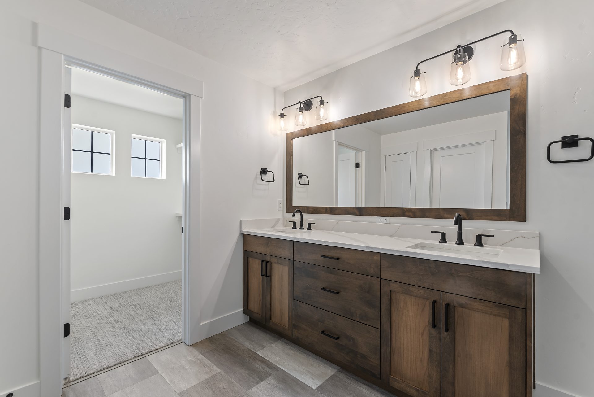 Bathroom featuring a double sink vanity with brown stain, a mirror matching the length of the vanity with coordinating stained trim, and double three-light fixtures. Includes one towel holder on each side of the vanity, mixed-color tile flooring, and an e