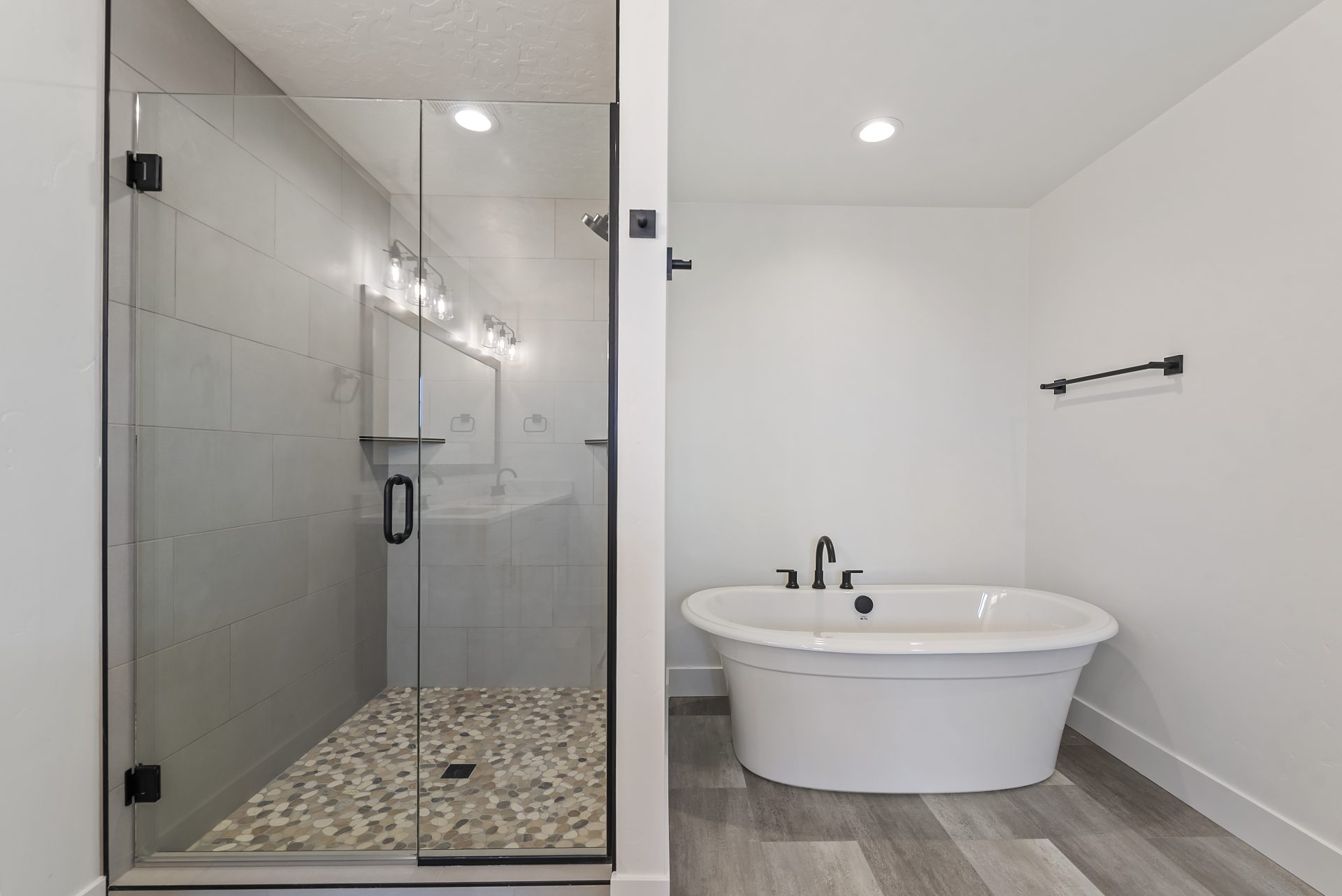 Bathroom featuring a freestanding tub, mixed gray and white tile flooring, a standing shower with natural rock floor, and a glass door with black finishes.