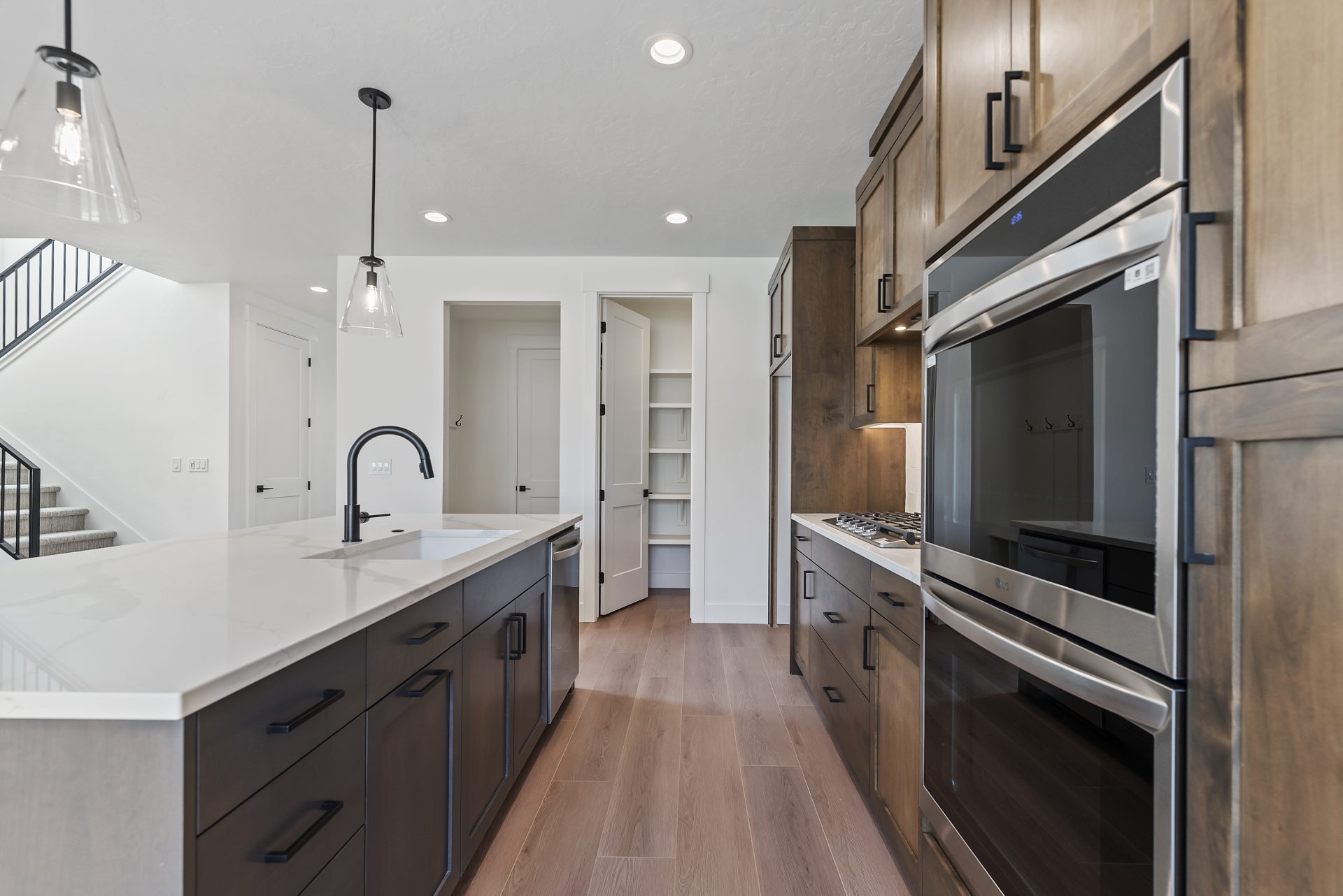 Kitchen viewed from an angle across the long island, featuring a double oven with gas range, abundant brown-stained cabinetry, medium brown wood flooring, and a view of the white shelved pantry with access to the mudroom.