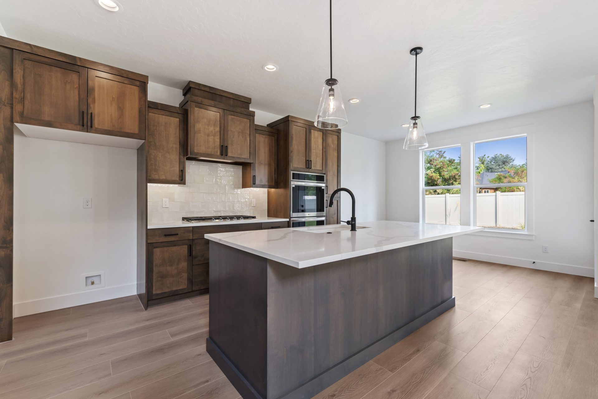 Kitchen viewed from the opposite side of the island, showing brown-stained cabinetry, a tile backsplash above the gas range, double oven, medium brown wood flooring, and an open space for the refrigerator (not shown)