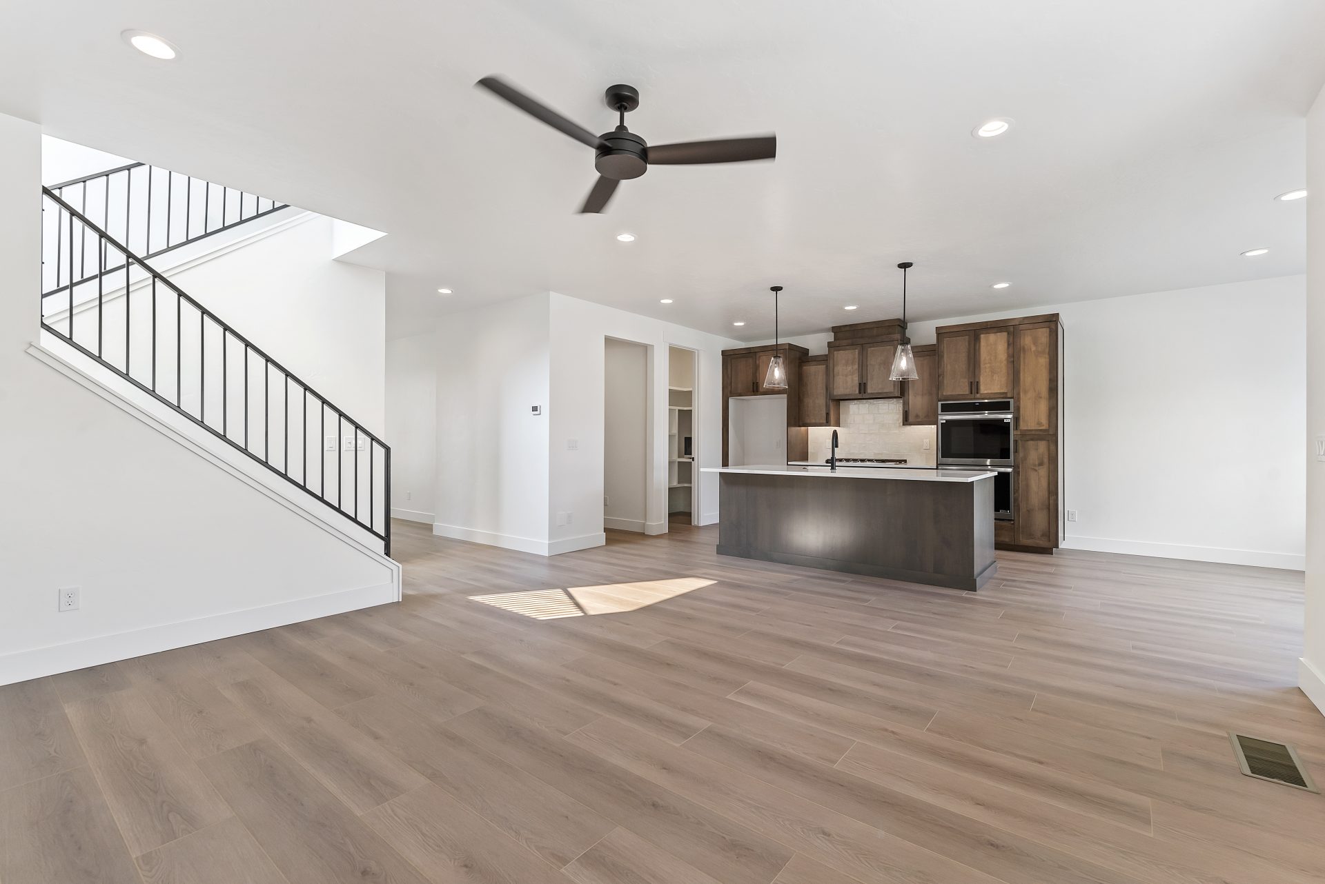 View from the living area across the room, showing medium brown wood flooring and the kitchen in the distance with brown-stained cabinetry and a long island.