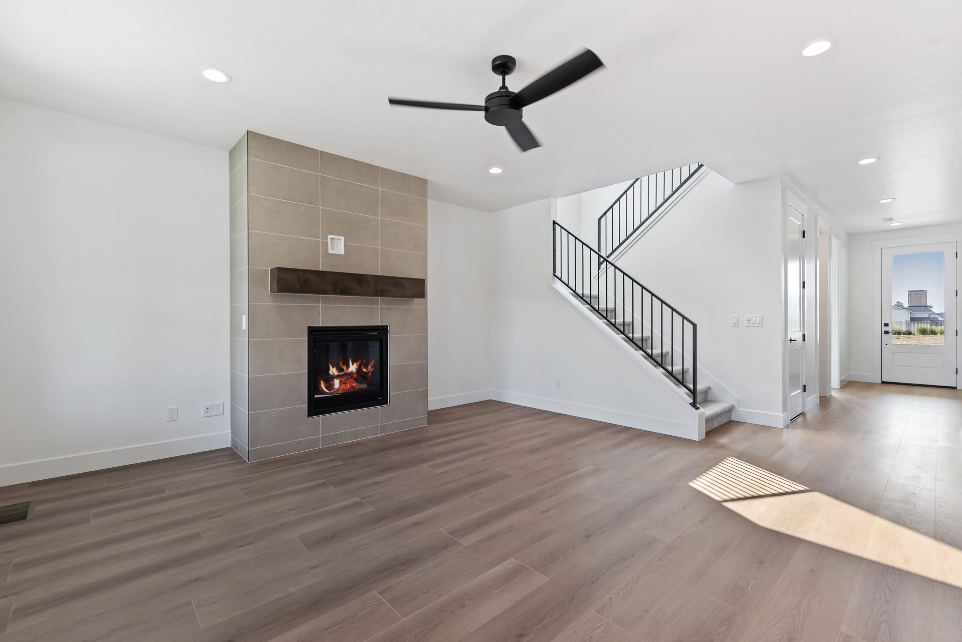 Living area viewed from across the room, featuring a fireplace with wall-to-ceiling gray tile and a brown-stained mantle, along with stairs and a window visible in the background
