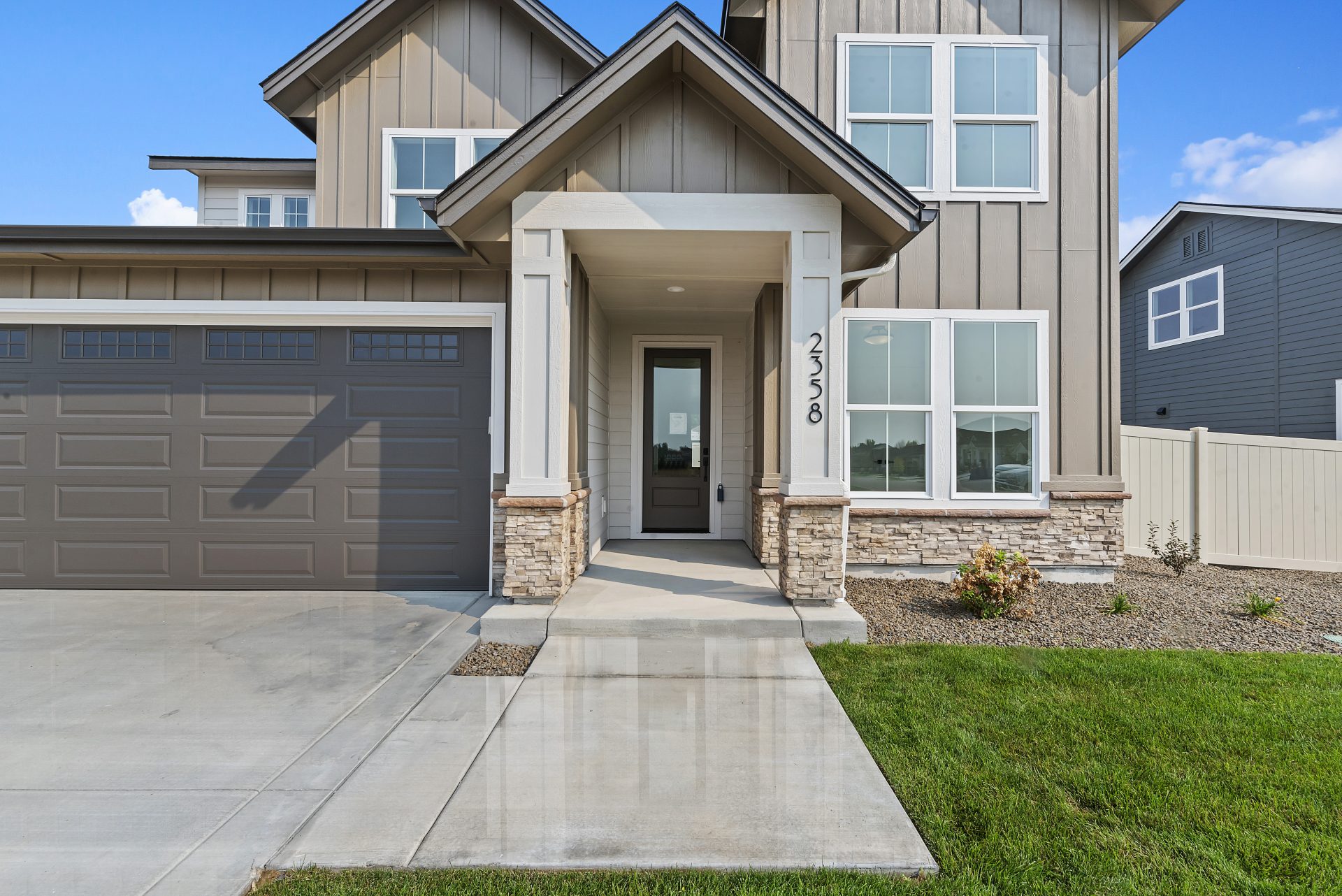Front exterior of a modern two-story house with tan siding, stone accents, a covered entryway, and a brown garage door. The walkway leads to the front door, and the yard features a small patch of grass and landscaping.