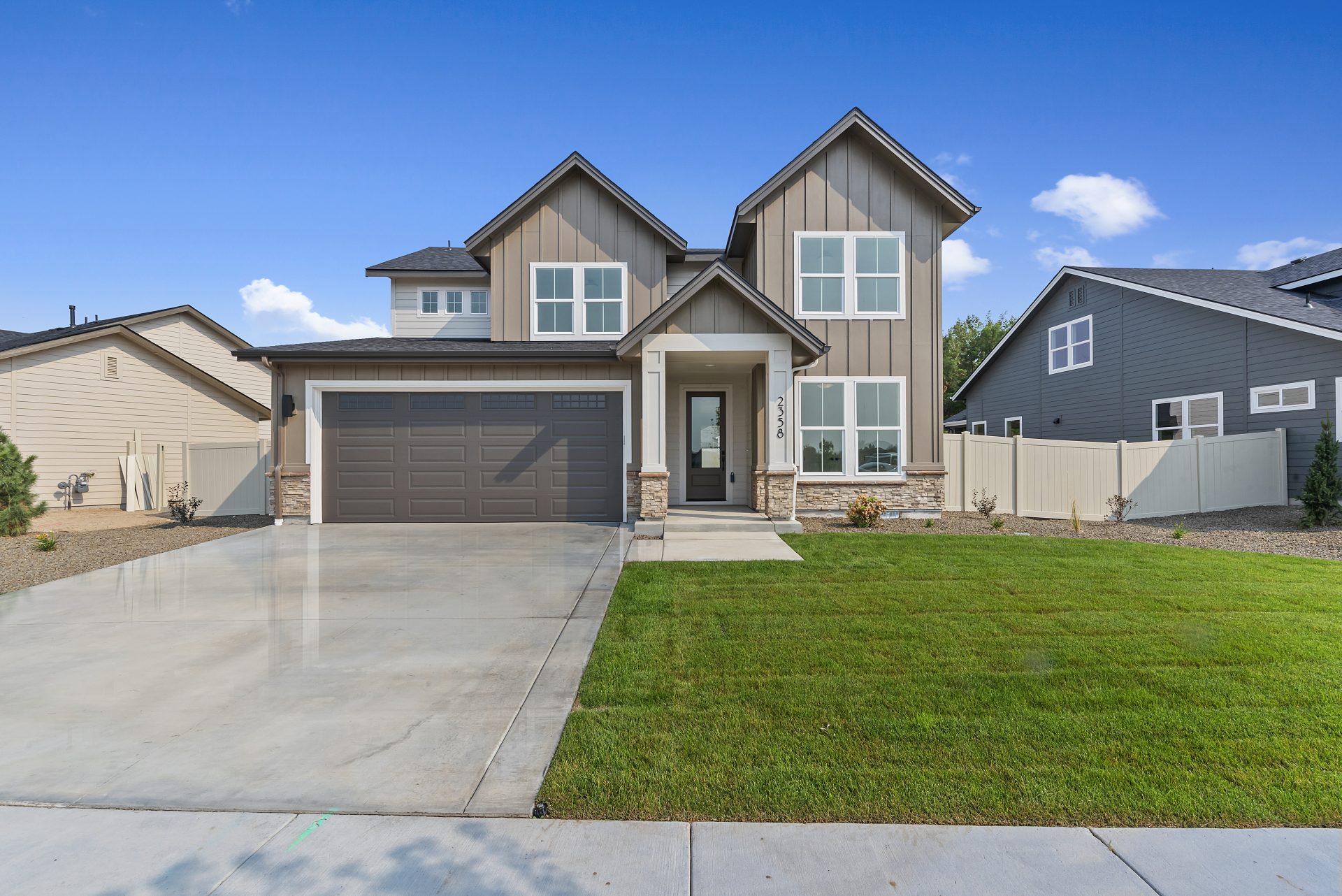 Front view of a modern two-story house with tan siding, stone accents, and a dark brown garage door. The home features multiple large windows, a covered entryway, and a neatly landscaped front yard with green grass and a wide driveway.
