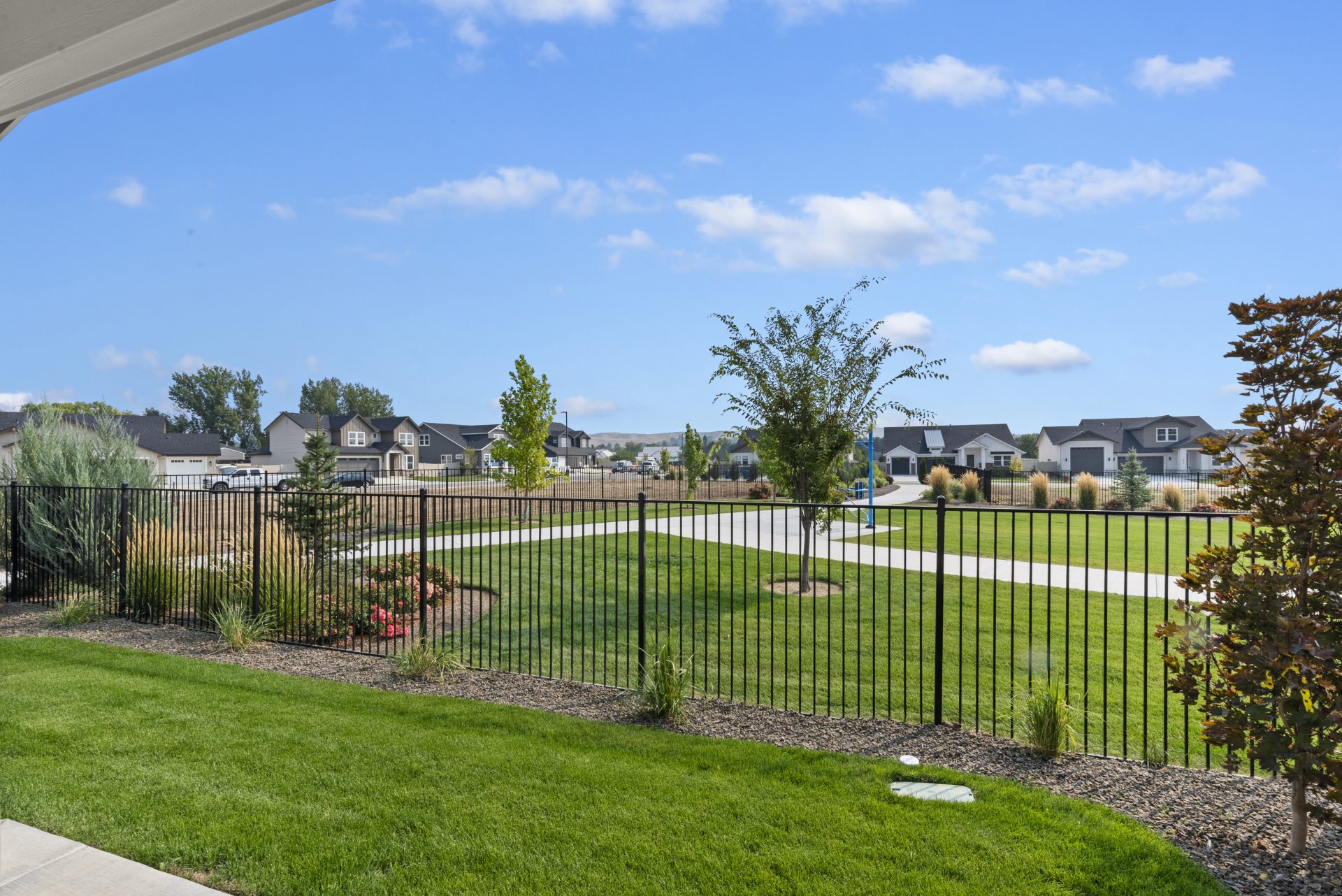 View of a landscaped backyard with green grass, flowerbeds, and a black iron fence, overlooking a neighborhood with trees and houses in the distance under a blue sky.