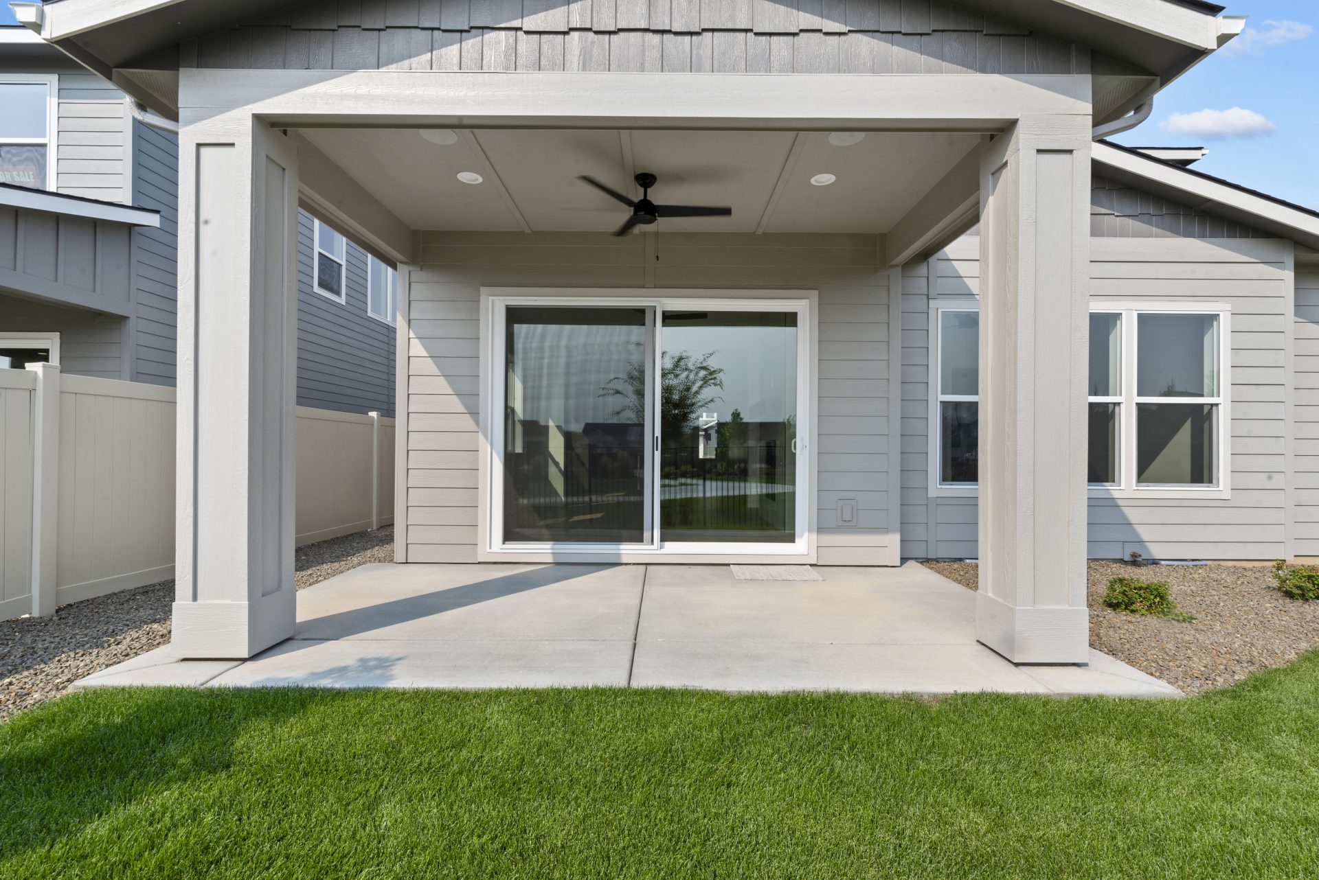 View of a modern home with covered patio including a ceiling fan, with a slider and windows on the right side of the rear of the home, with grass and landscaped plants and rock.