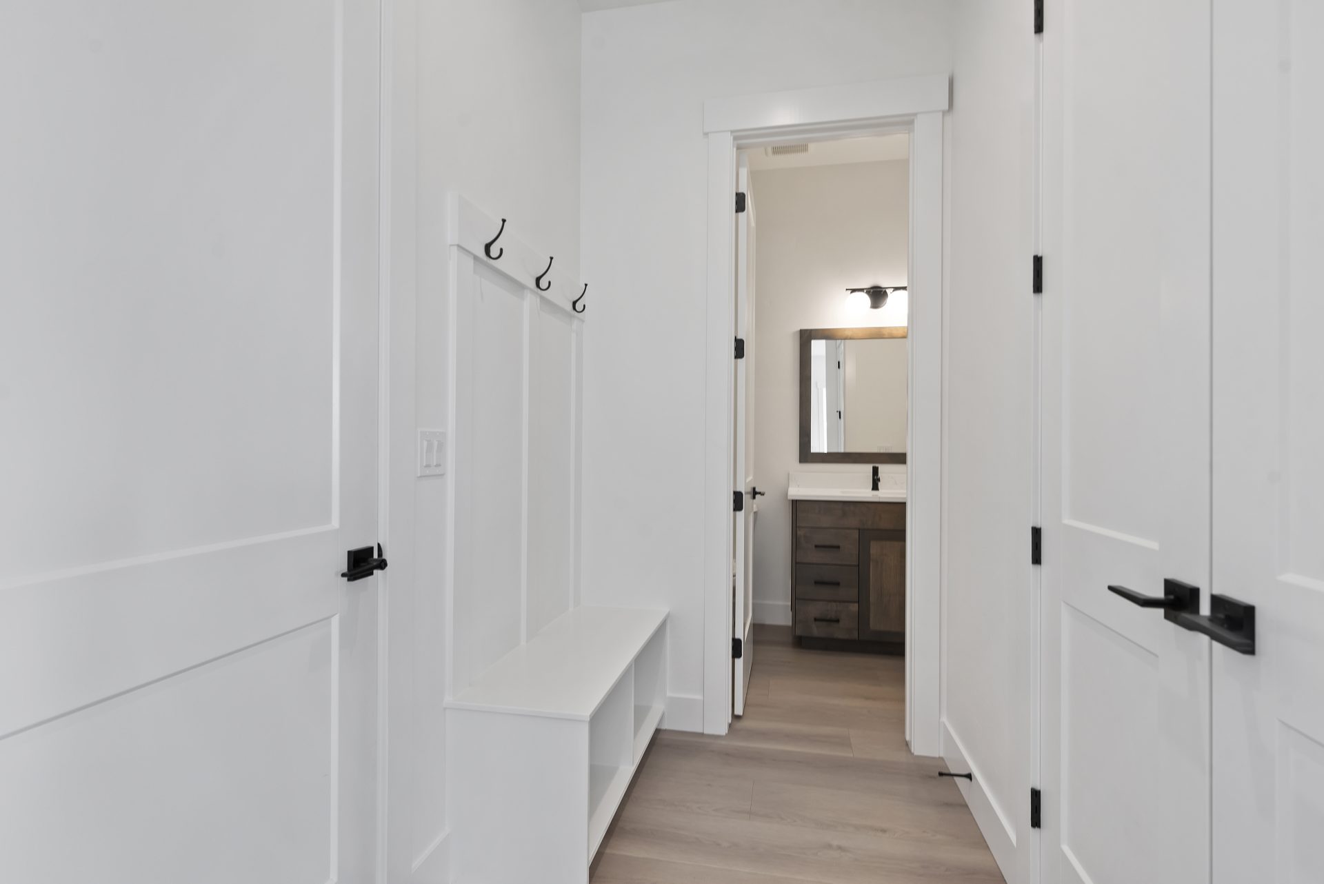 View of a room with all white walls, white paneling with a bench and storage cubbies underneath, hooks above the paneling located above the bench seat and an entry door to a bathroom with a vanity.